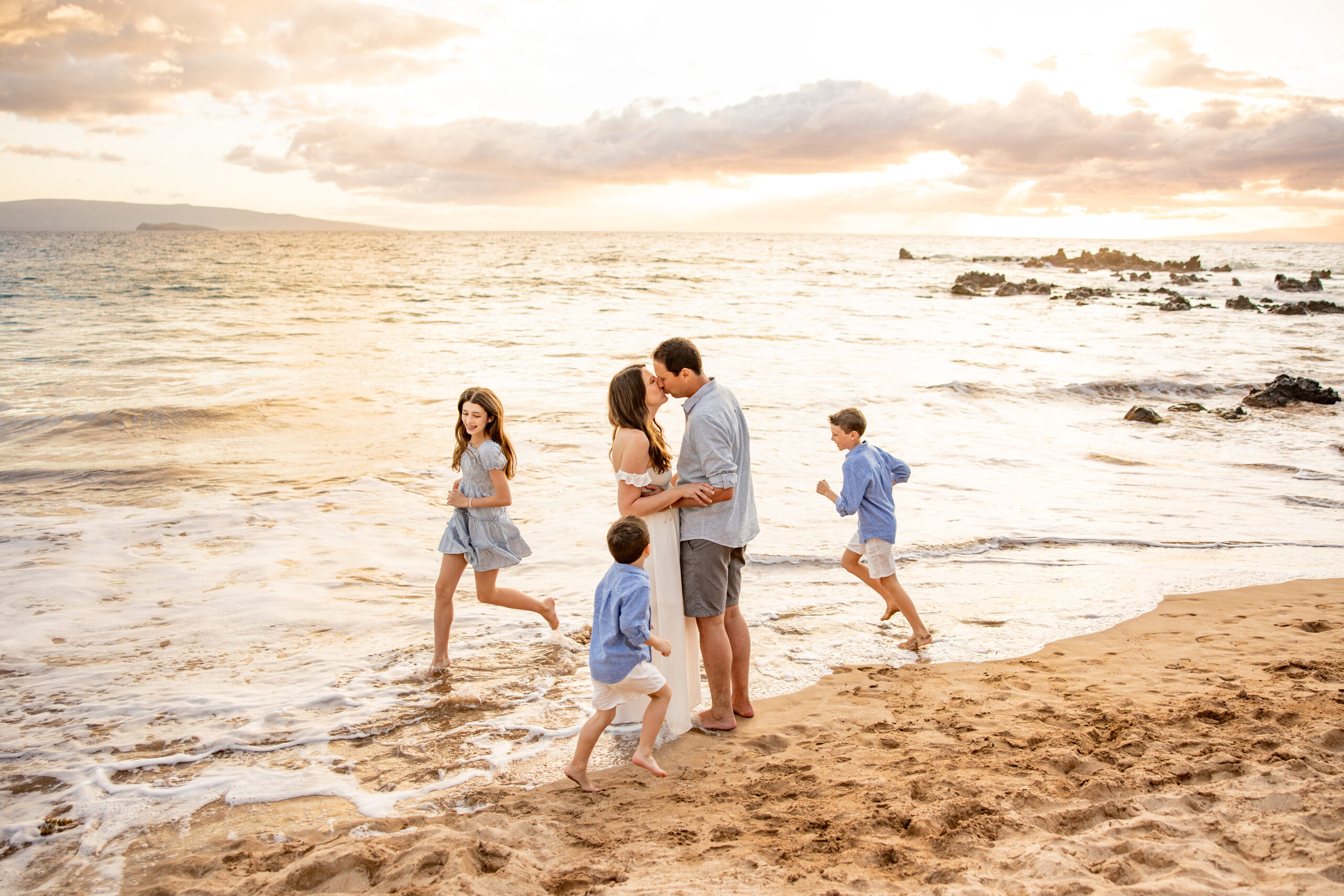 Maui Family Photographer captures mother and father kissing while children run around on the beach during Maui family photos