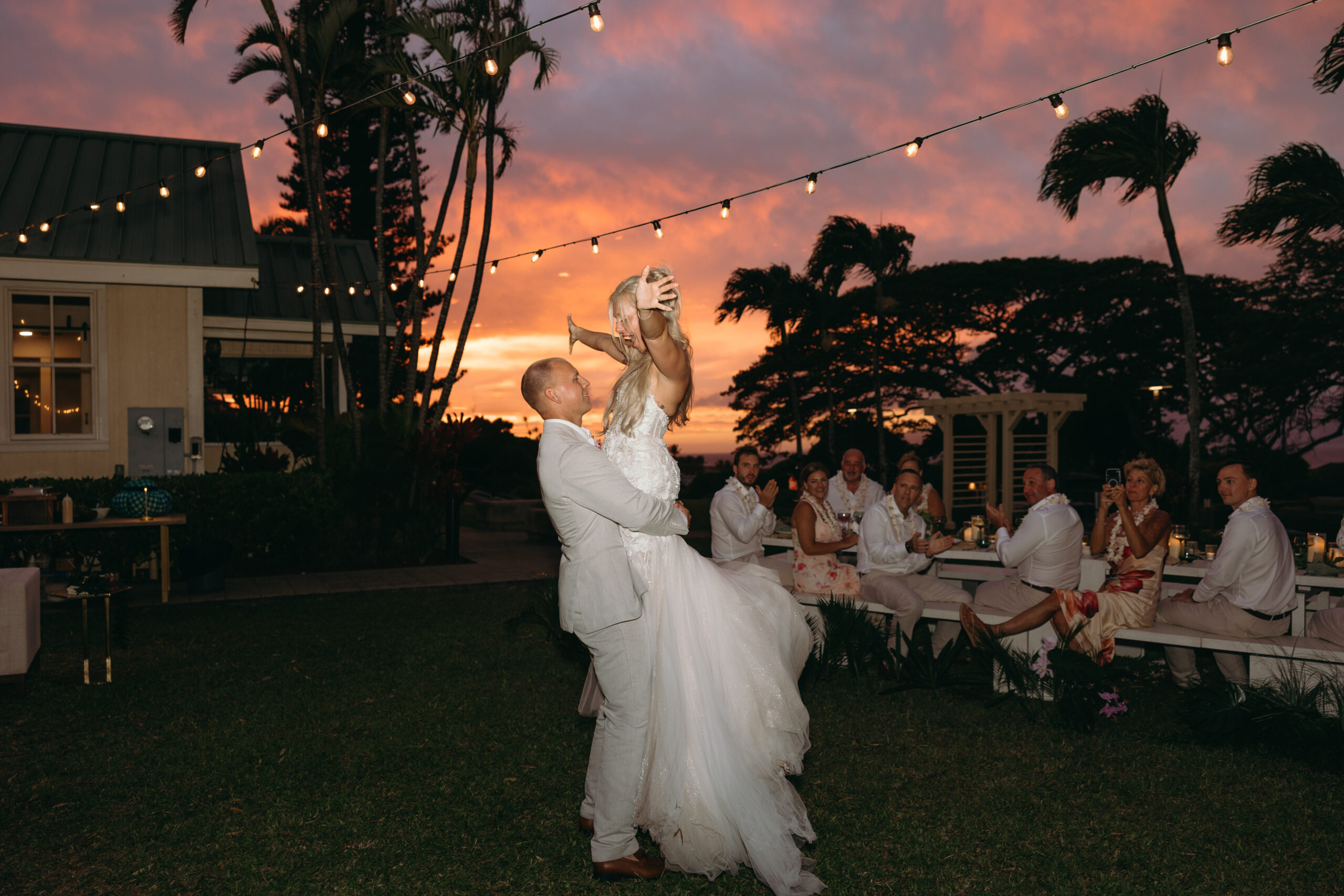 A bride twirls with joy as her groom lifts her in the air under glowing string lights and a stunning sunset sky—pure Hawaii wedding planning magic.