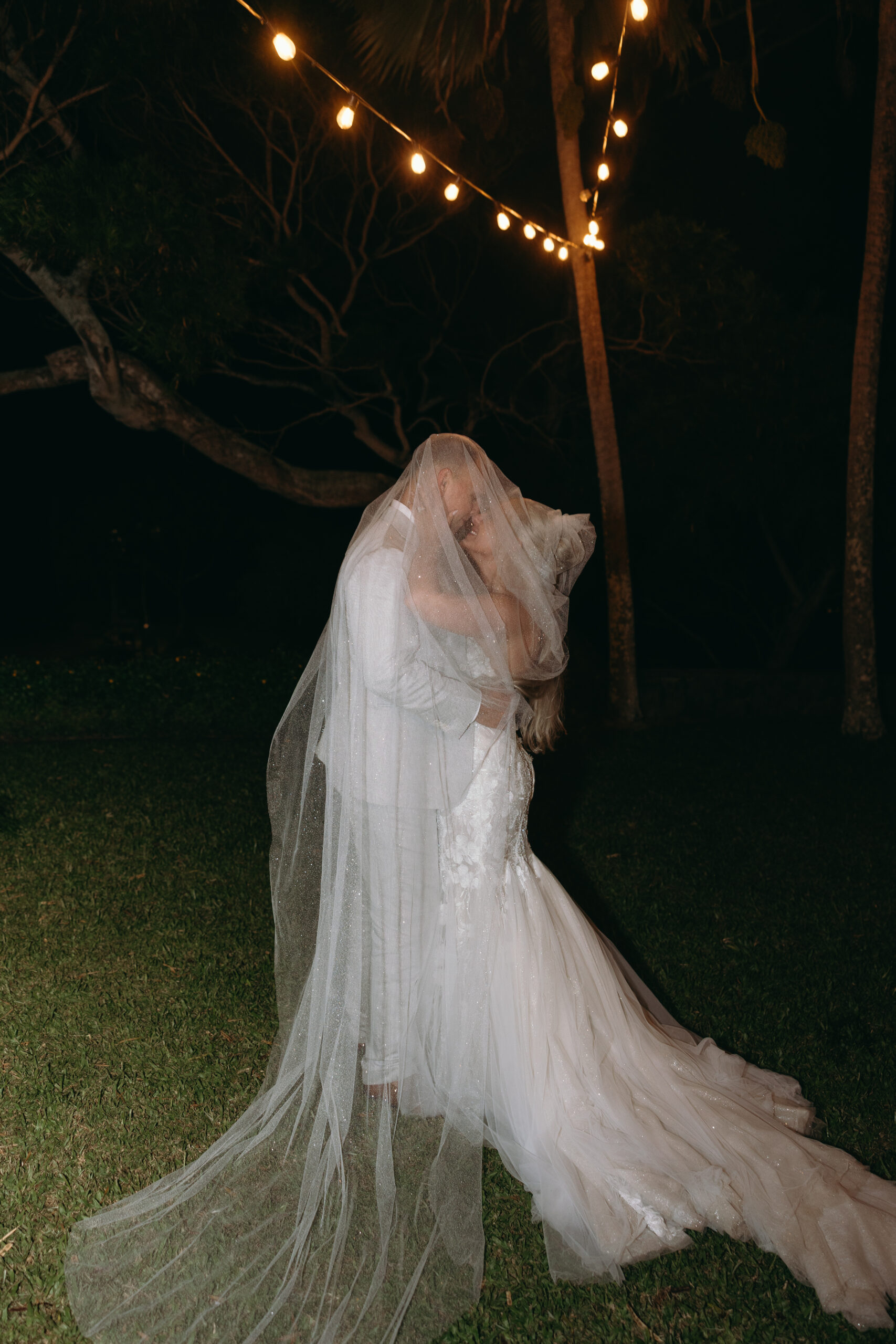 A couple shares a tender nighttime embrace under glowing string lights, wrapped in her veil and the magic of their Hawaii wedding day.