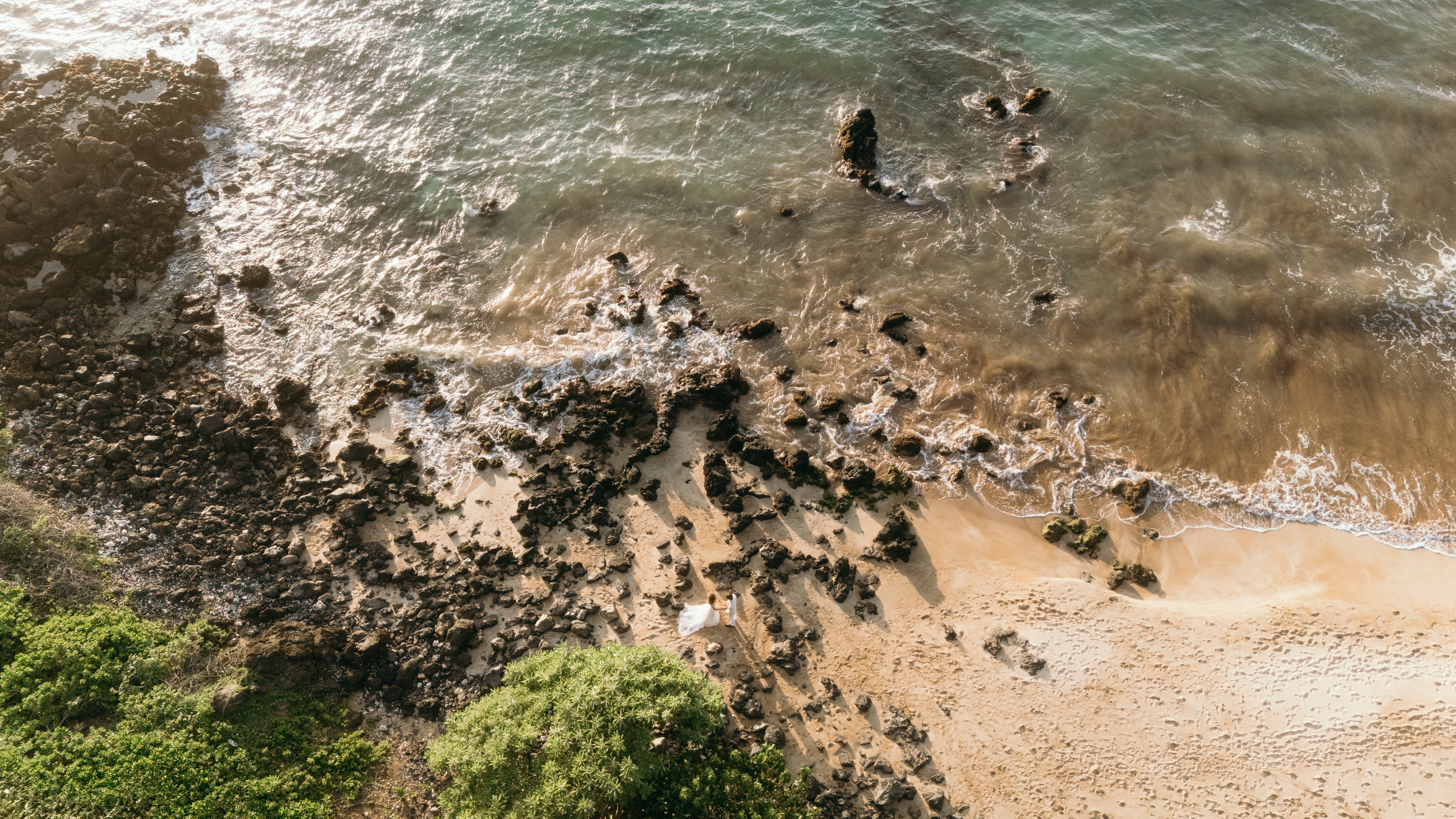 Aerial view of a newlywed couple walking hand in hand along a rocky Maui shoreline, the golden light and soft waves creating a magical Hawaii wedding planning moment.