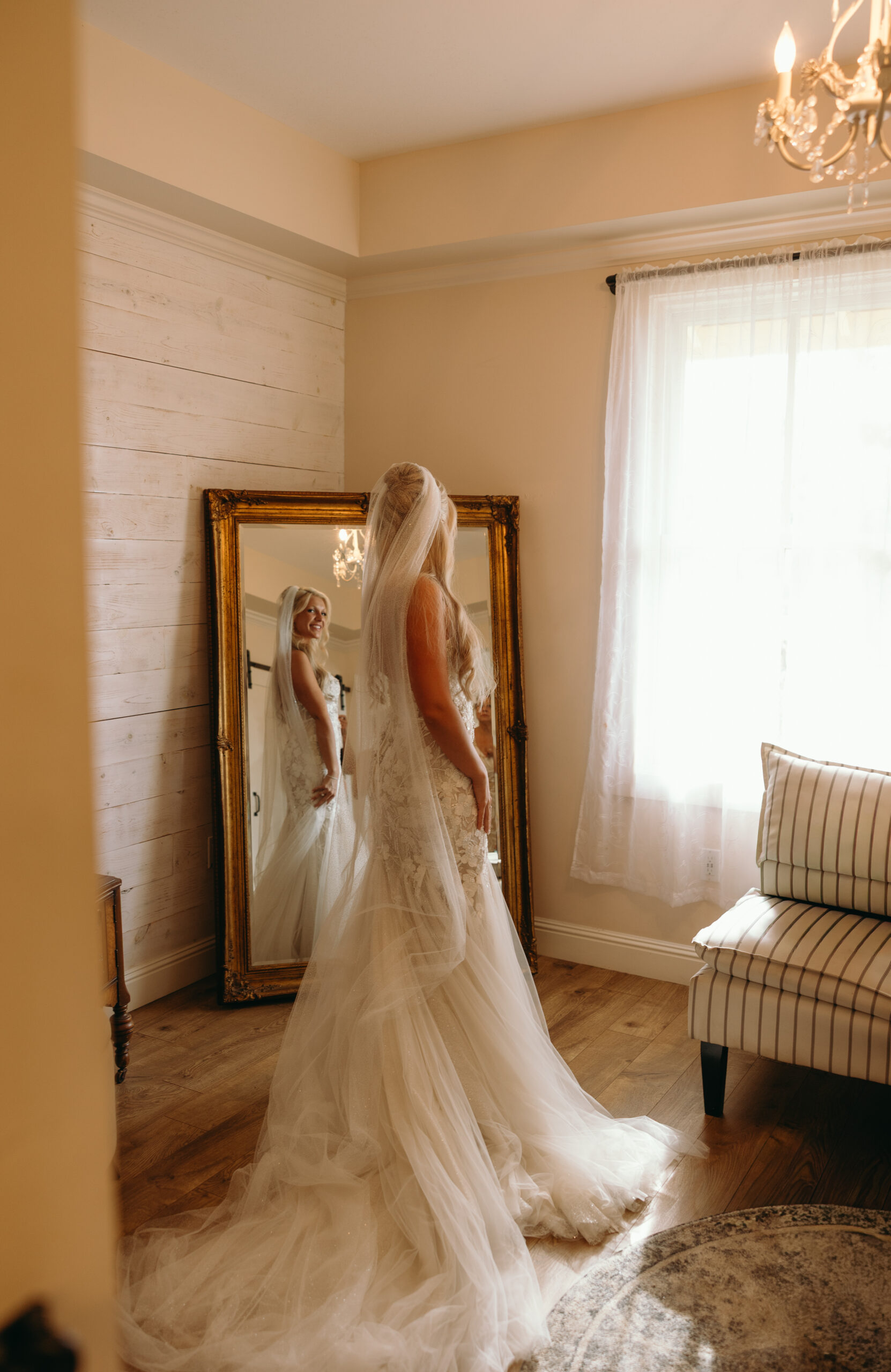 A bride gazes at her reflection in a tall gold-framed mirror, her veil and train cascading down the floor in soft light.