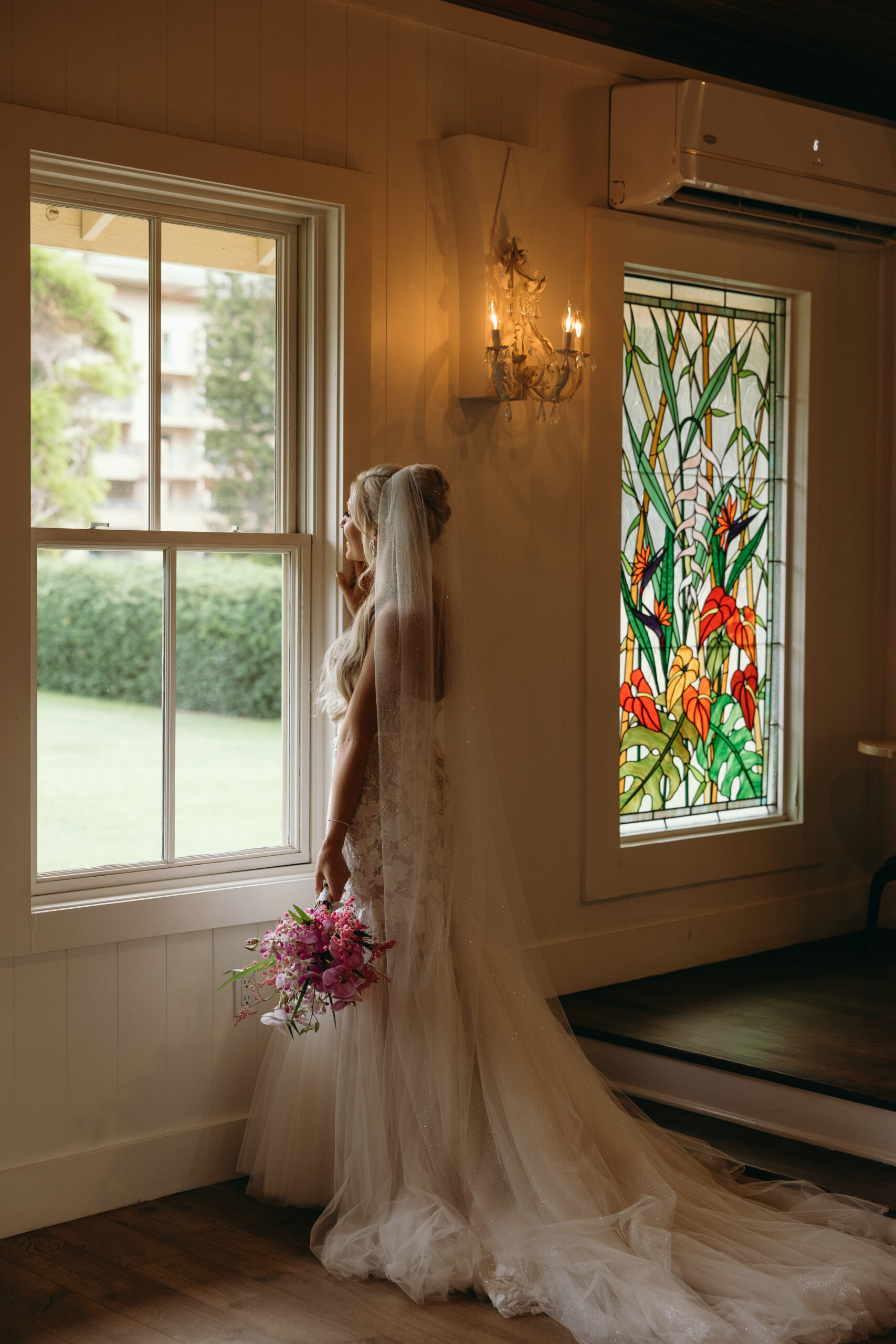 A bride stands quietly at a window, bouquet in hand, surrounded by soft stained glass and warm wooden walls—one of many beautiful moments made possible through Hawaii wedding planning.