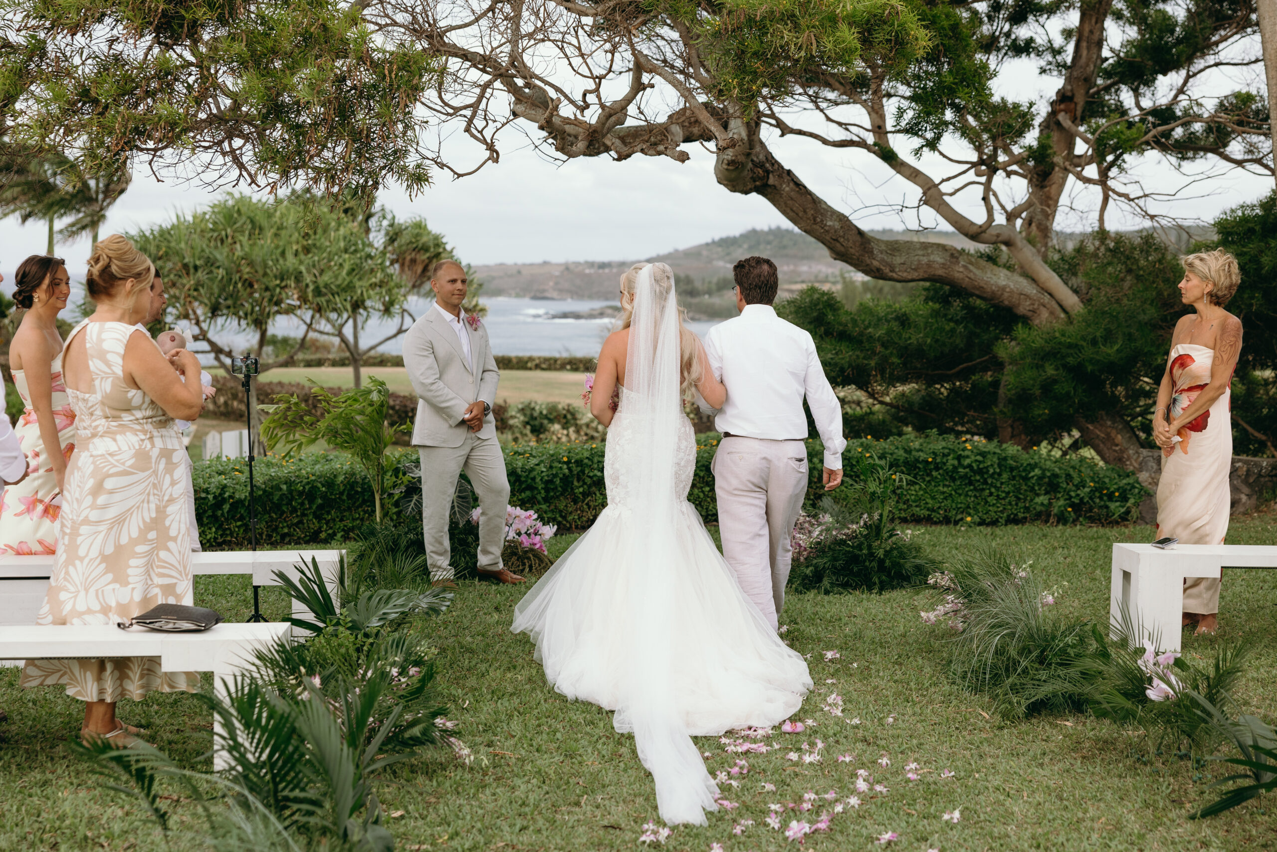 A couple exchanges vows under a wooden arch draped in florals and surrounded by palm trees, with loved ones looking on in celebration.