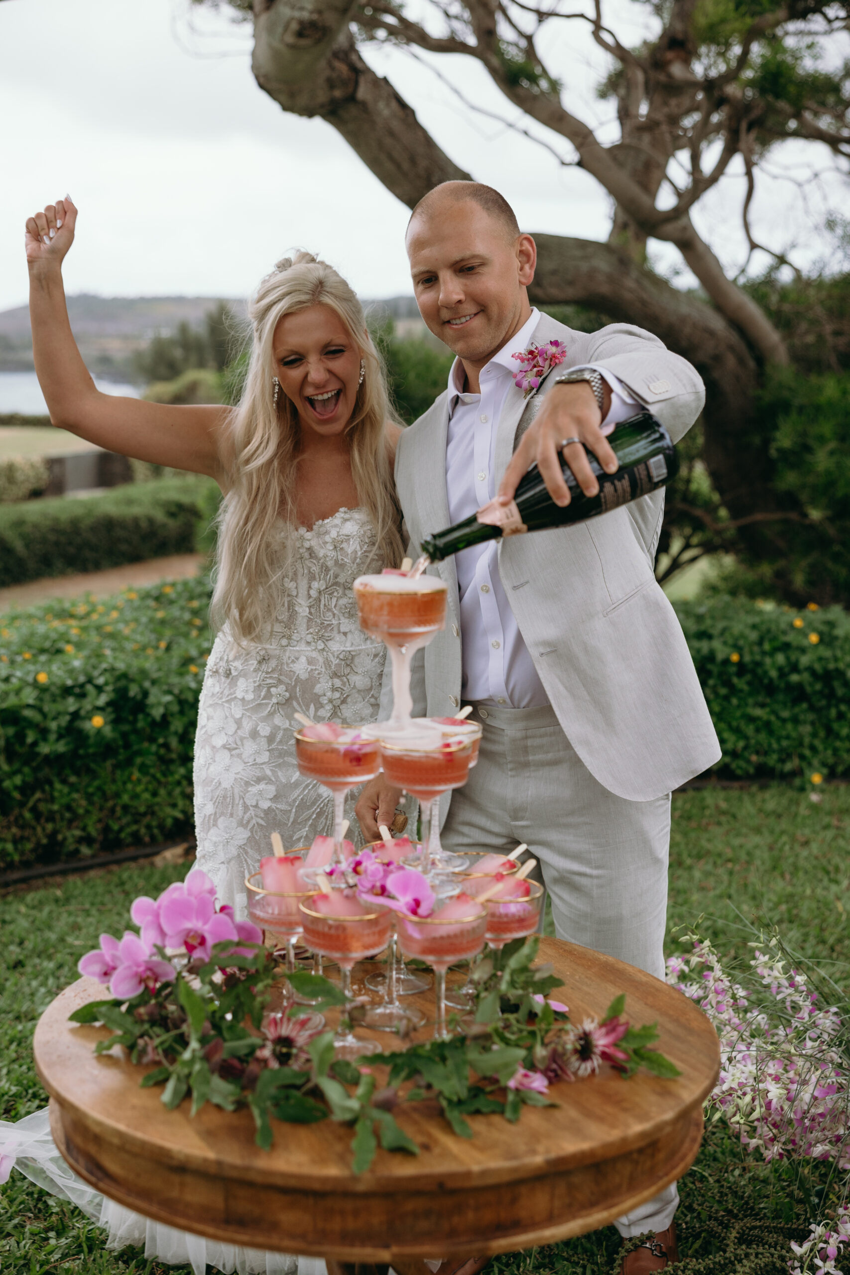 A groom pours sparkling rosé into a tower of coupe glasses while the bride cheers beside him—Hawaii wedding planning with a fun and festive twist.