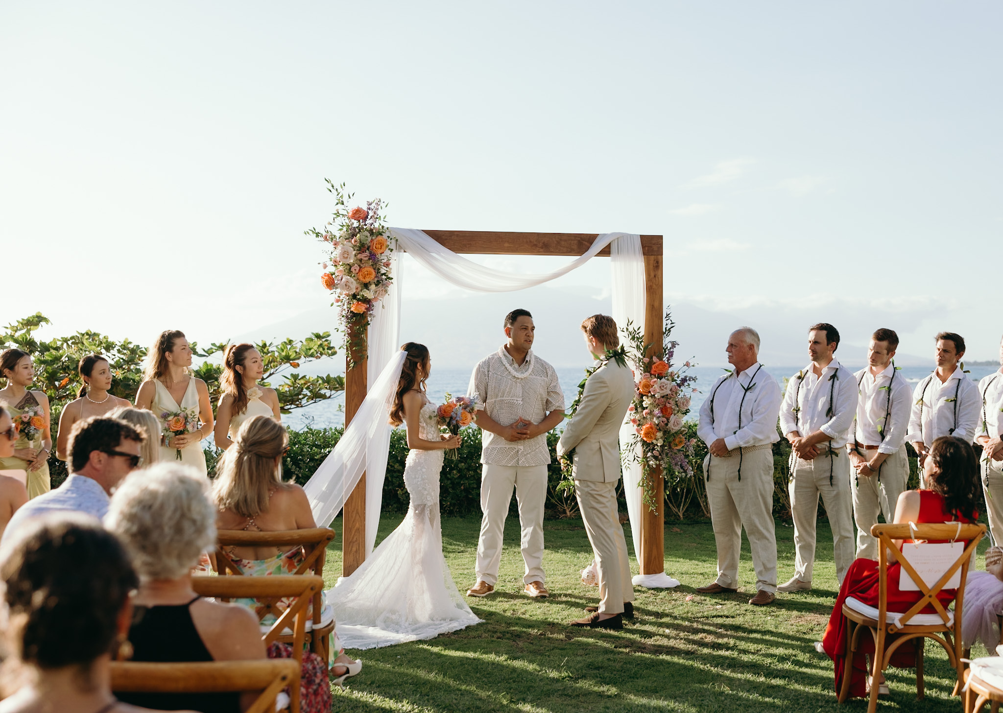 A couple exchanges vows under a floral arch with their wedding party looking on, Maui’s ocean and mountains providing the perfect backdrop for their Hawaii wedding planning vision.