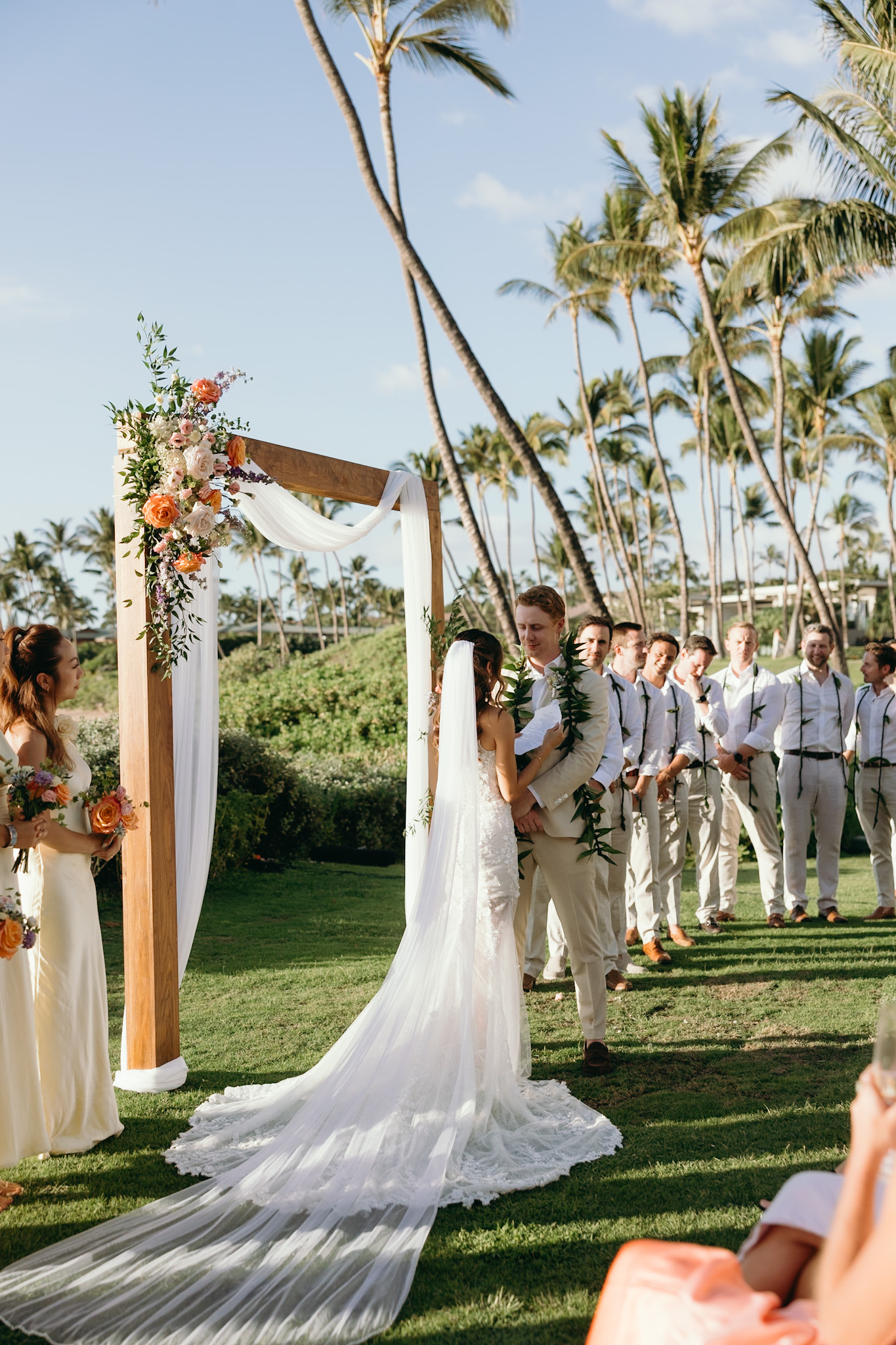 A couple exchanges vows under a wooden arch draped in florals and surrounded by palm trees, with loved ones looking on in celebration.