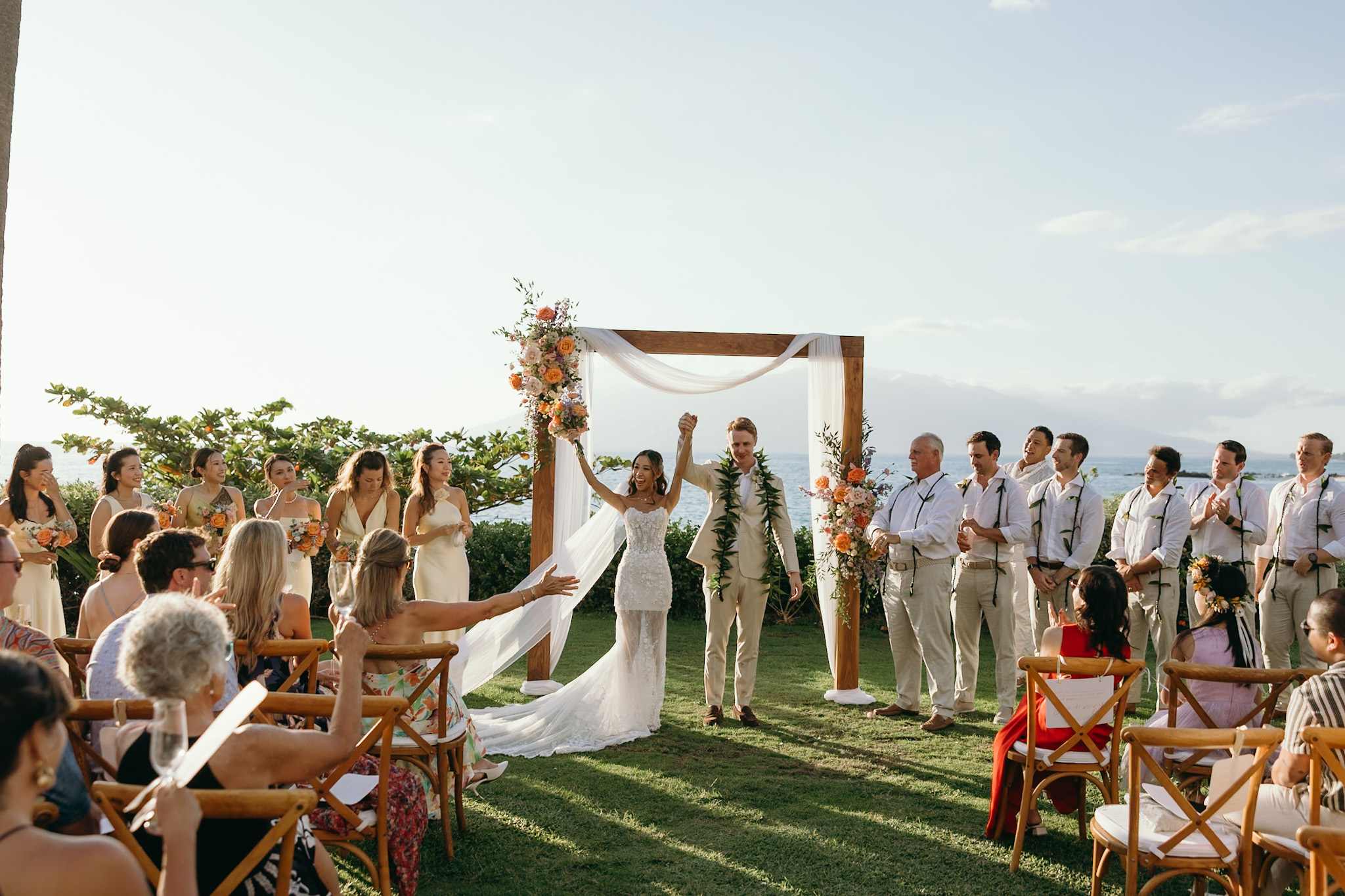 A bride and groom joyfully raise their hands under a floral ceremony arch in front of family and friends during their Maui wedding celebration.