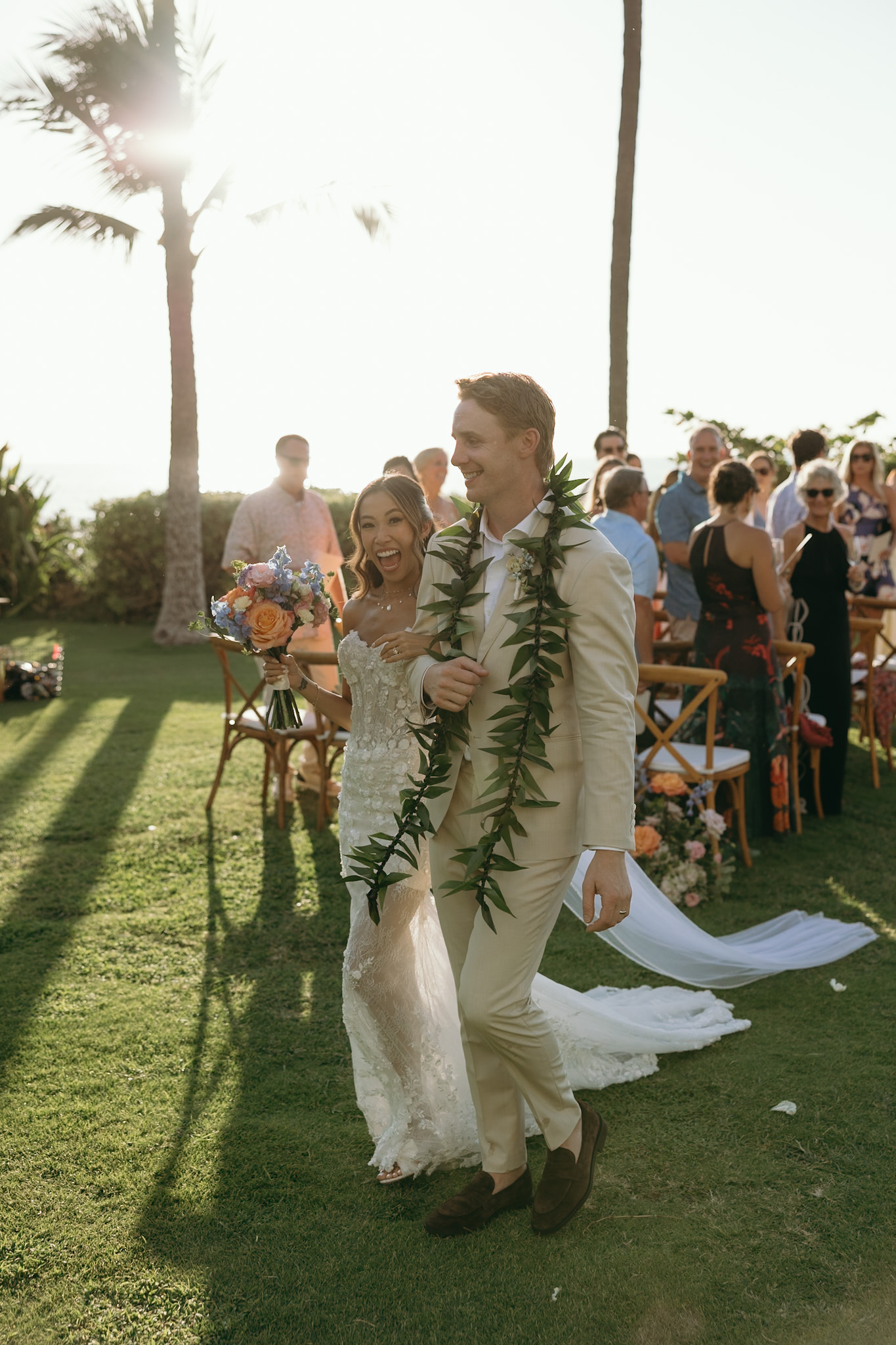 A newlywed couple walks joyfully down the aisle as guests cheer them on, the golden Maui sun glowing behind—Hawaii wedding planning dreams come true.