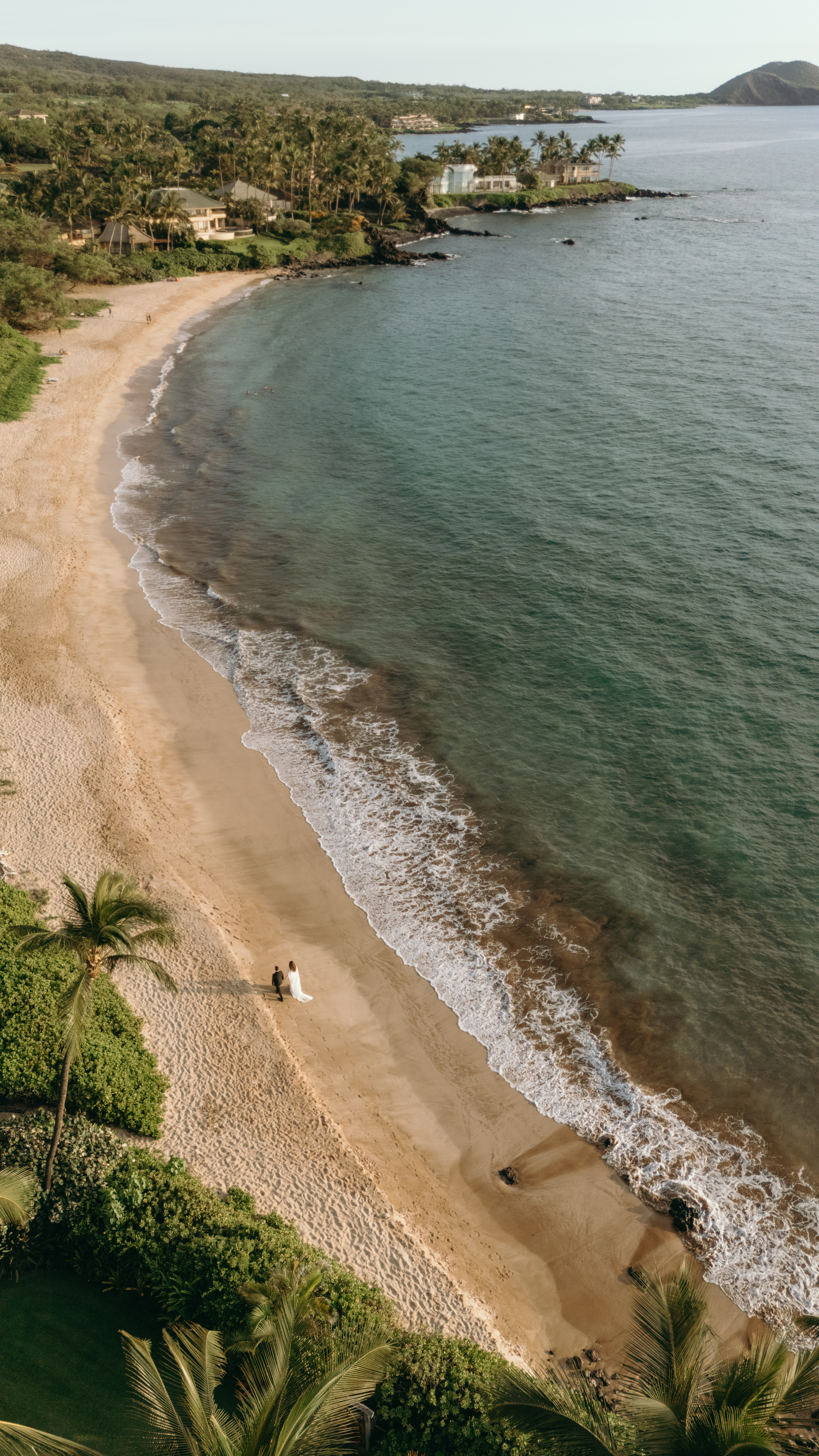 Drone shot of couple walking a curved shoreline on a clear, sunny Maui day