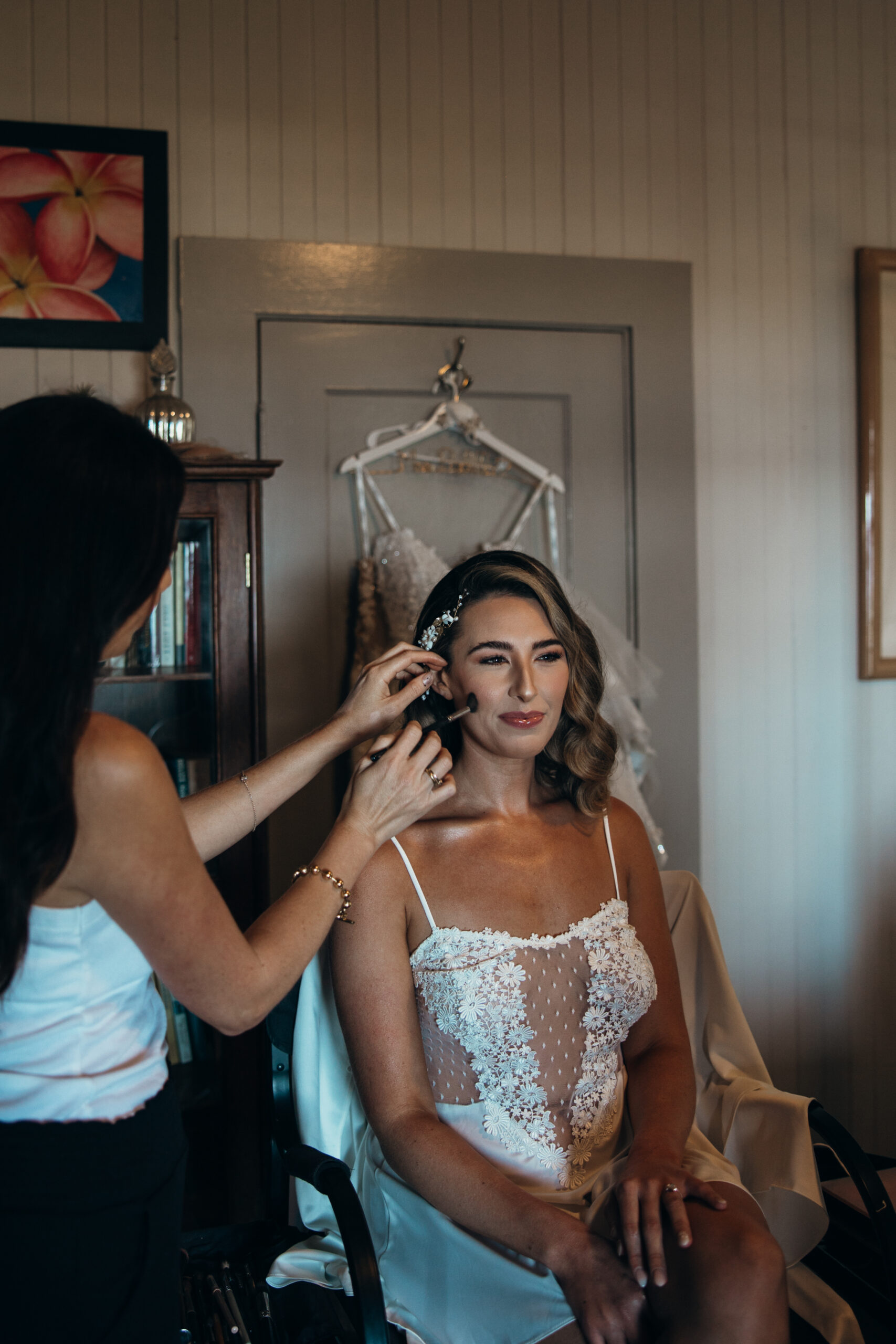 Bride getting her makeup touched up in a softly lit room, with her wedding dress hanging behind her.