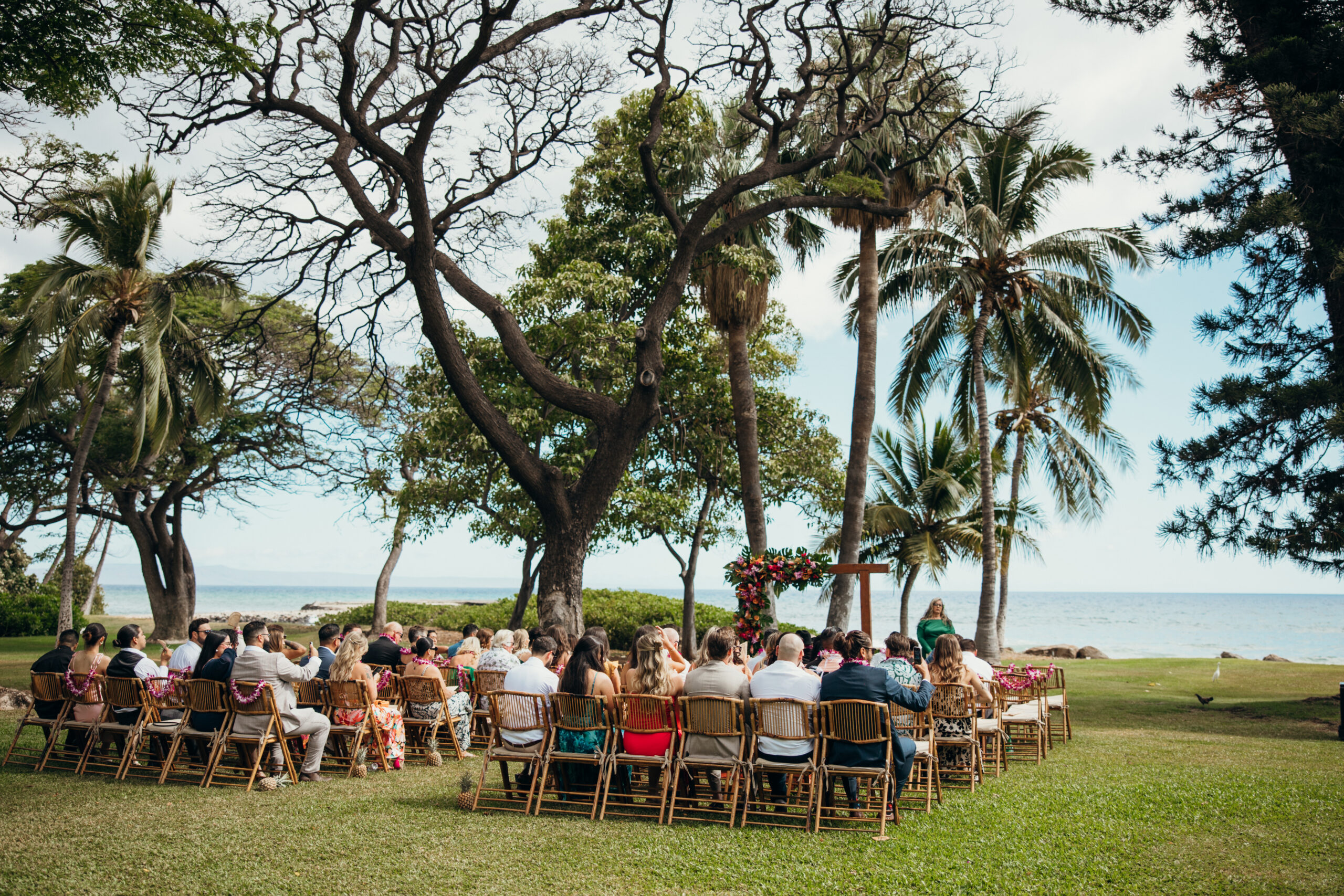 Guests seated under shady trees, facing the floral arch and ocean backdrop as the ceremony begins at a beachfront Maui wedding venue.