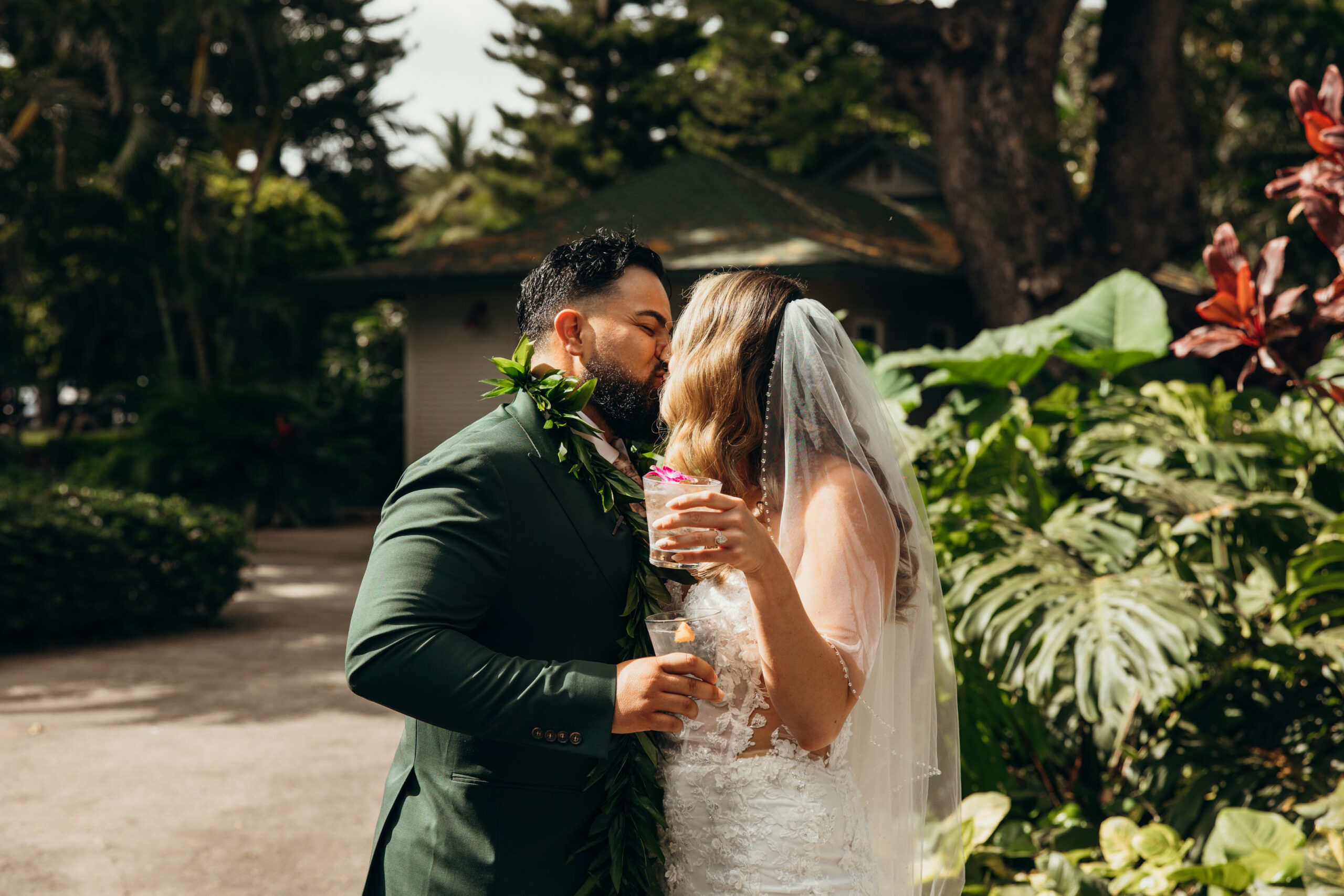 Bride and groom steal a moment with cocktails in hand, kissing beneath the tropical greenery during their Maui celebration.
