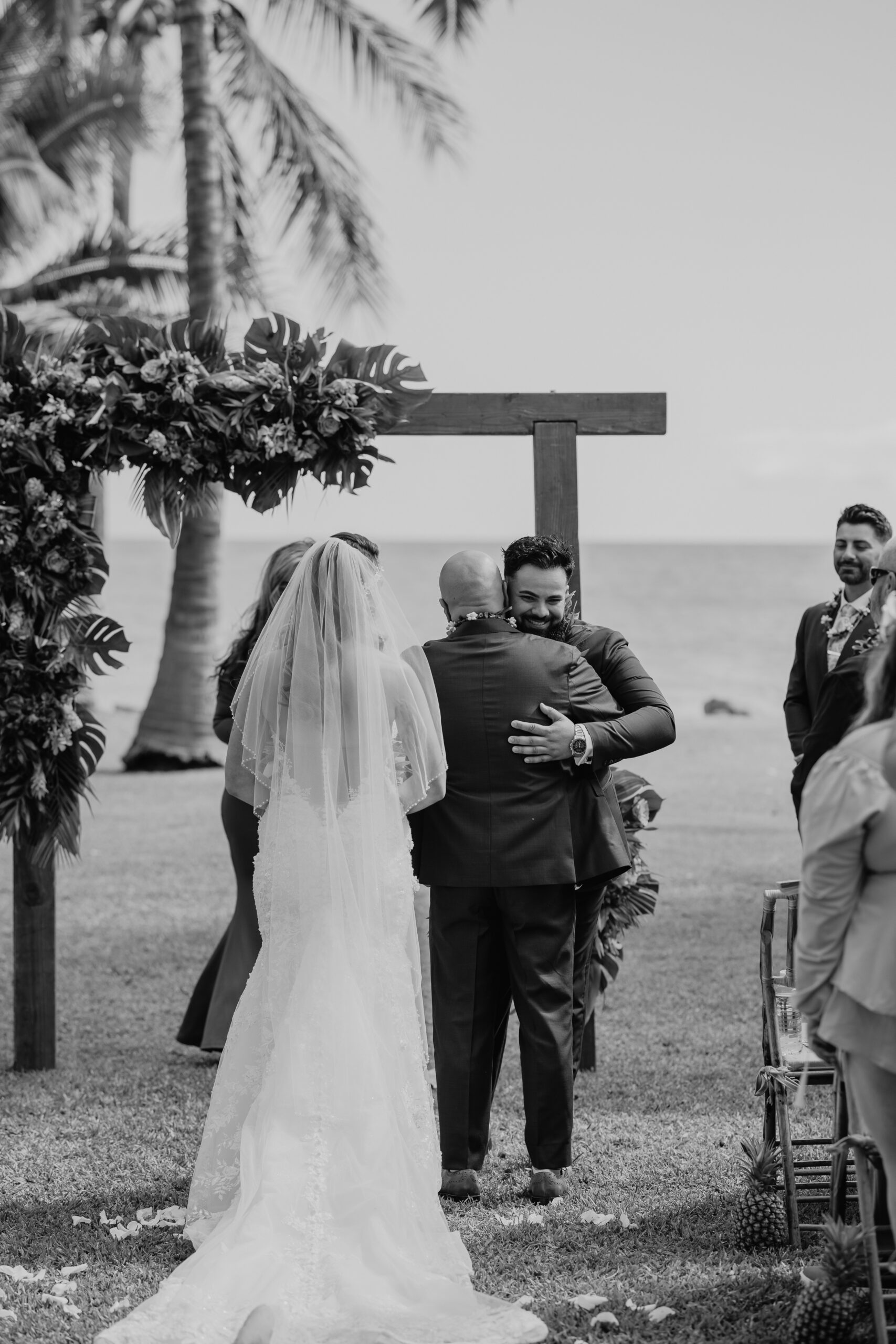 Groom hugs the bride’s father at the altar while the bride approaches, a heartfelt moment beneath the palm trees.