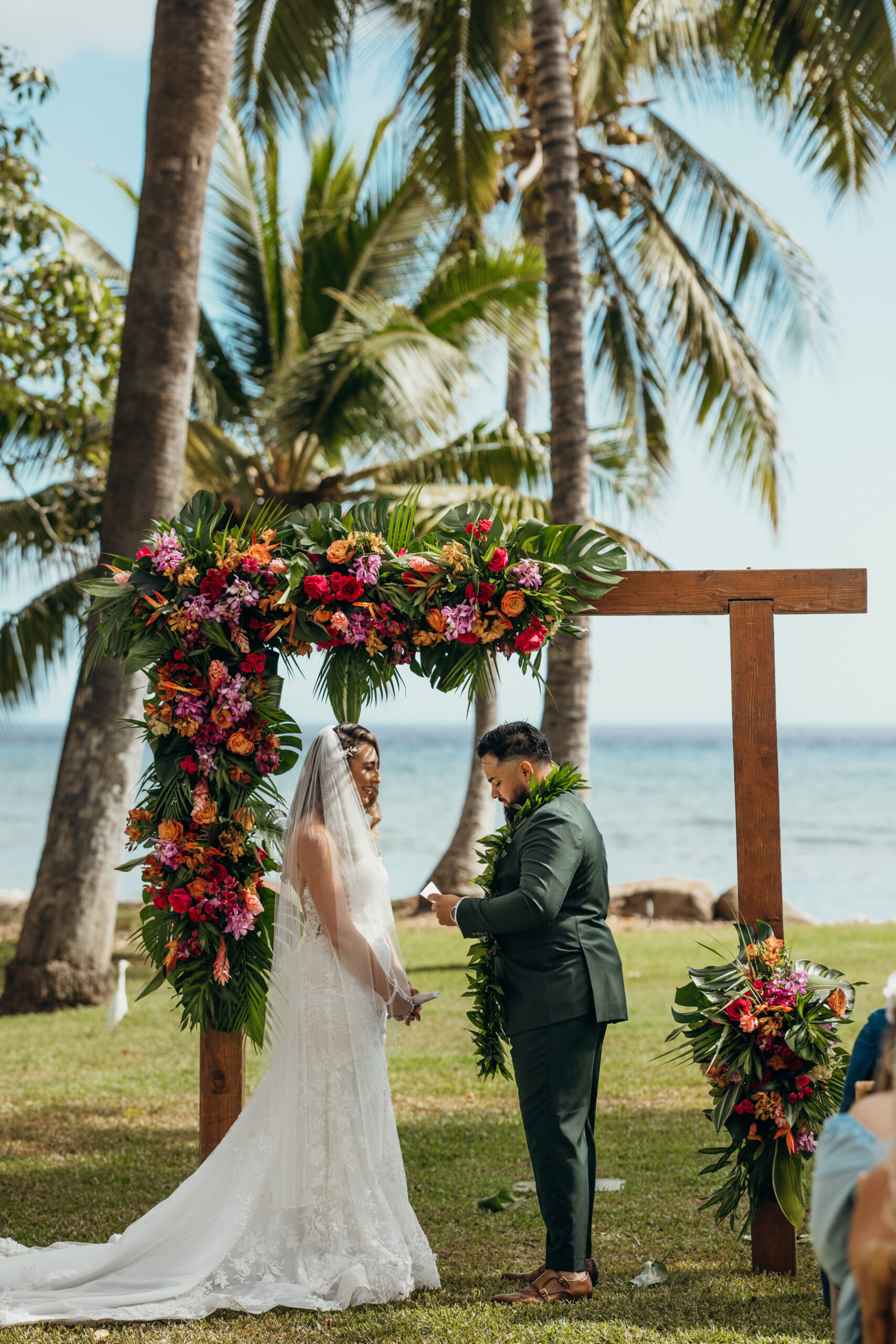 Groom places the ring on his bride’s finger beneath a lush floral arch at their oceanfront Maui wedding ceremony.