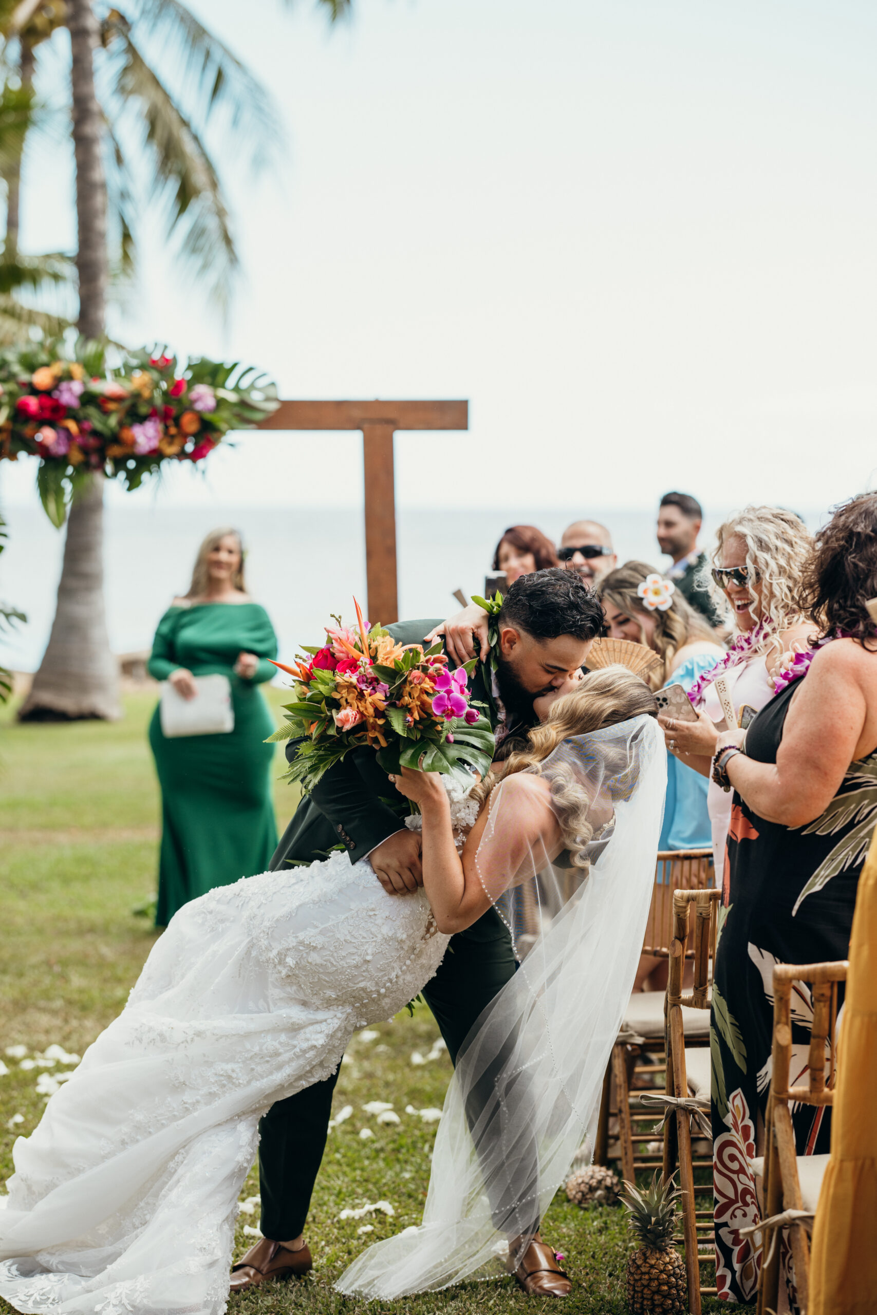 Groom dips the bride for a romantic post-ceremony kiss, surrounded by loved ones and island florals at their Maui wedding venue.