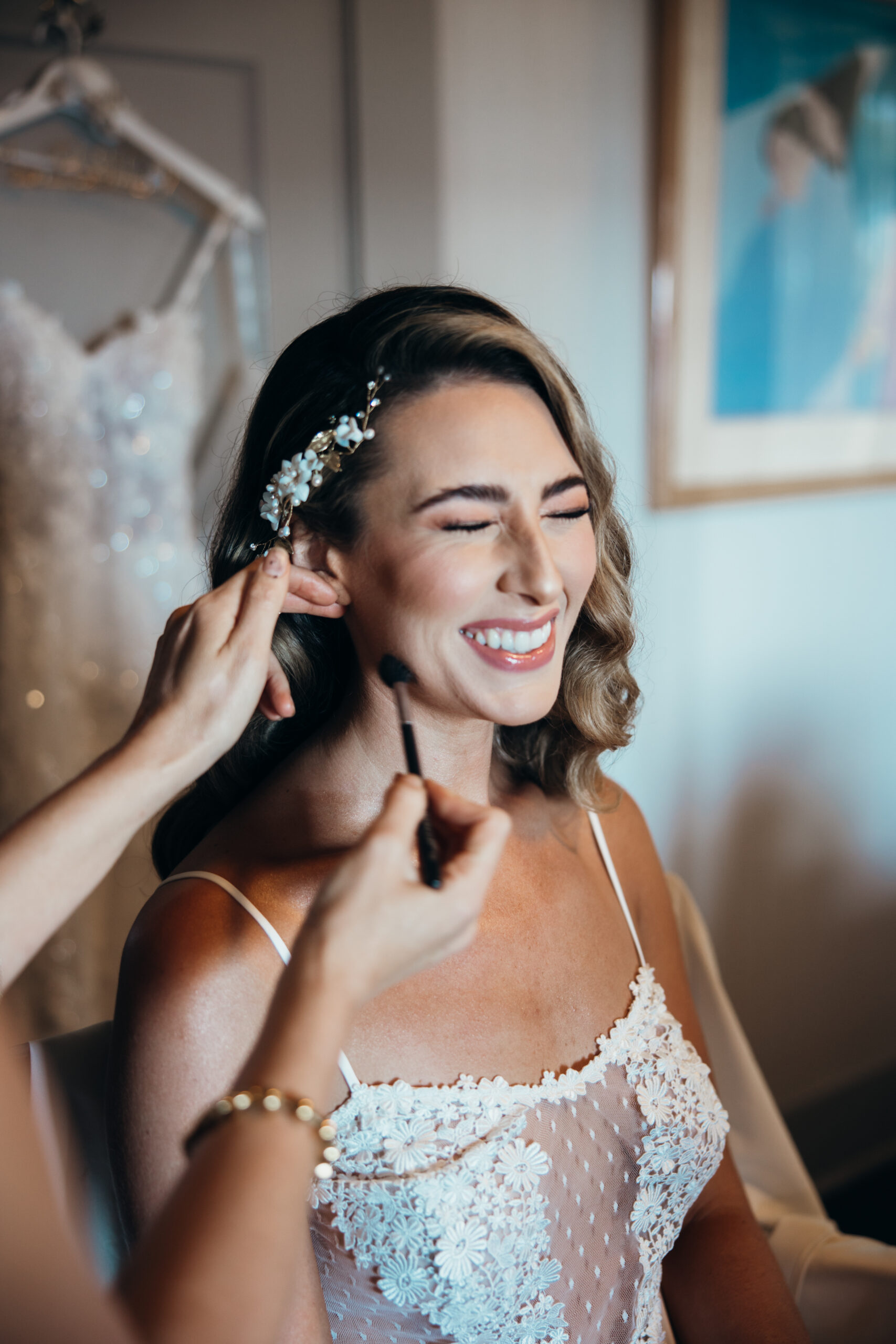 Bride smiling with eyes closed as she gets blush applied, wearing a delicate bridal robe while getting ready for her big day.