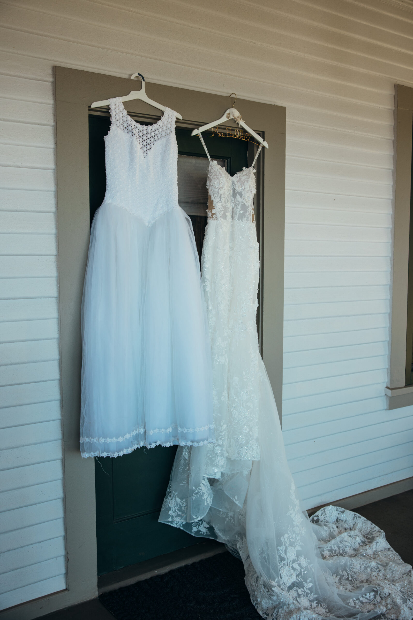 Two wedding dresses—one bridal gown and one child’s dress—hanging side-by-side outside a historic-style building at a Maui wedding venue.