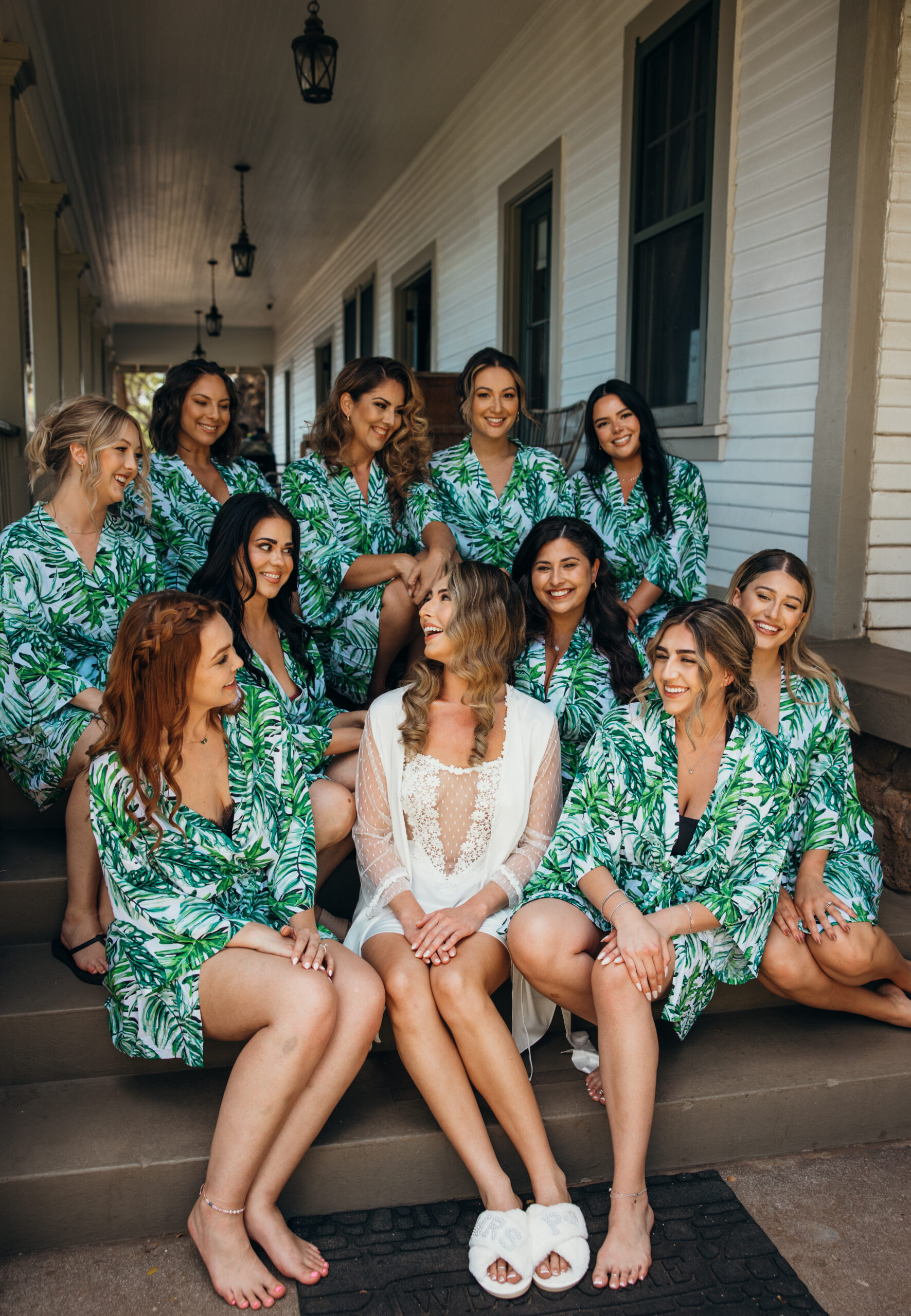 Bride sitting with her bridesmaids in matching palm leaf robes, sharing laughs on the steps of a plantation-style porch in Maui.
