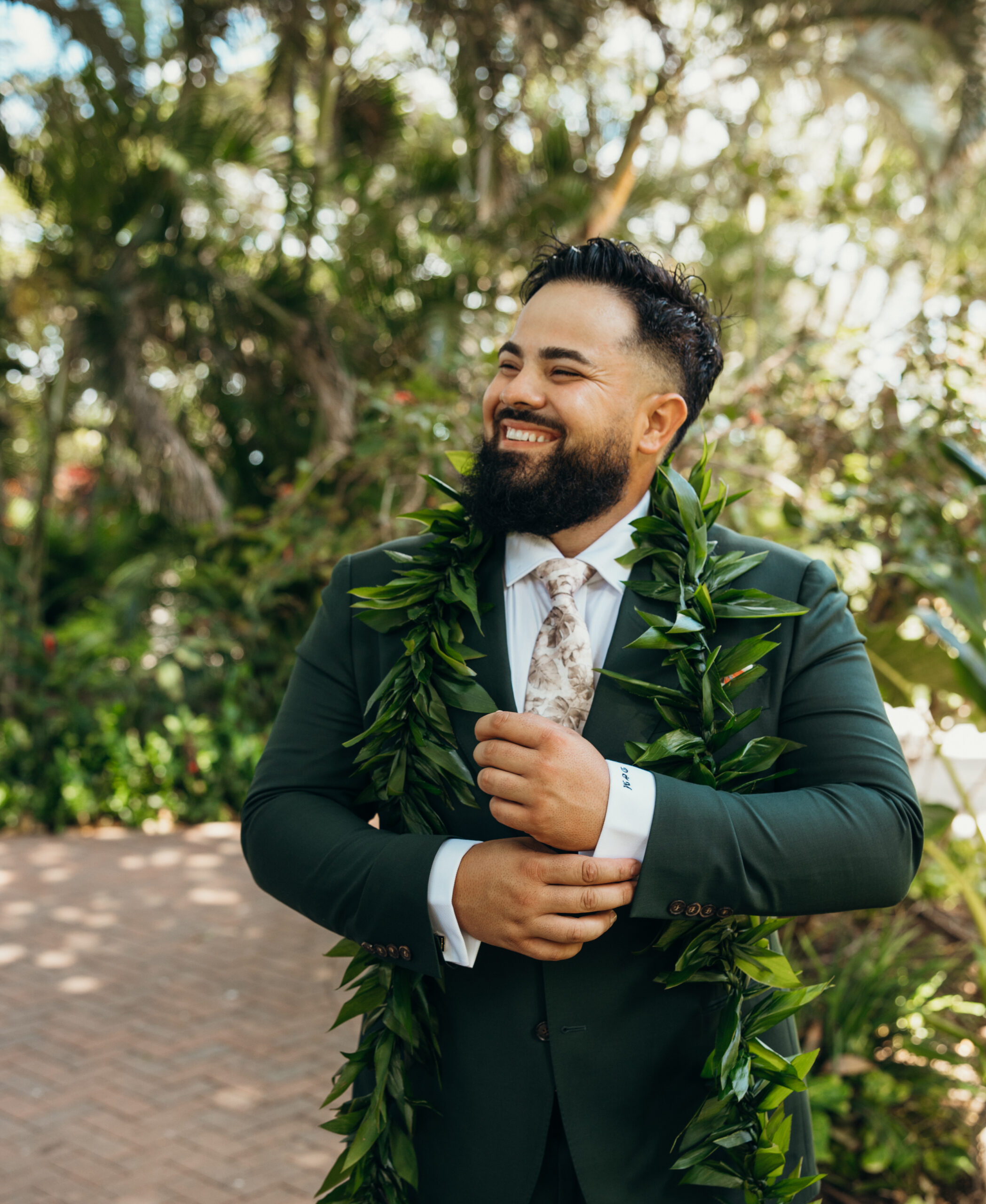 Groom smiling wide as he adjusts his sleeve, wearing a maile lei and tropical tie, surrounded by lush greenery at a Maui wedding venue.