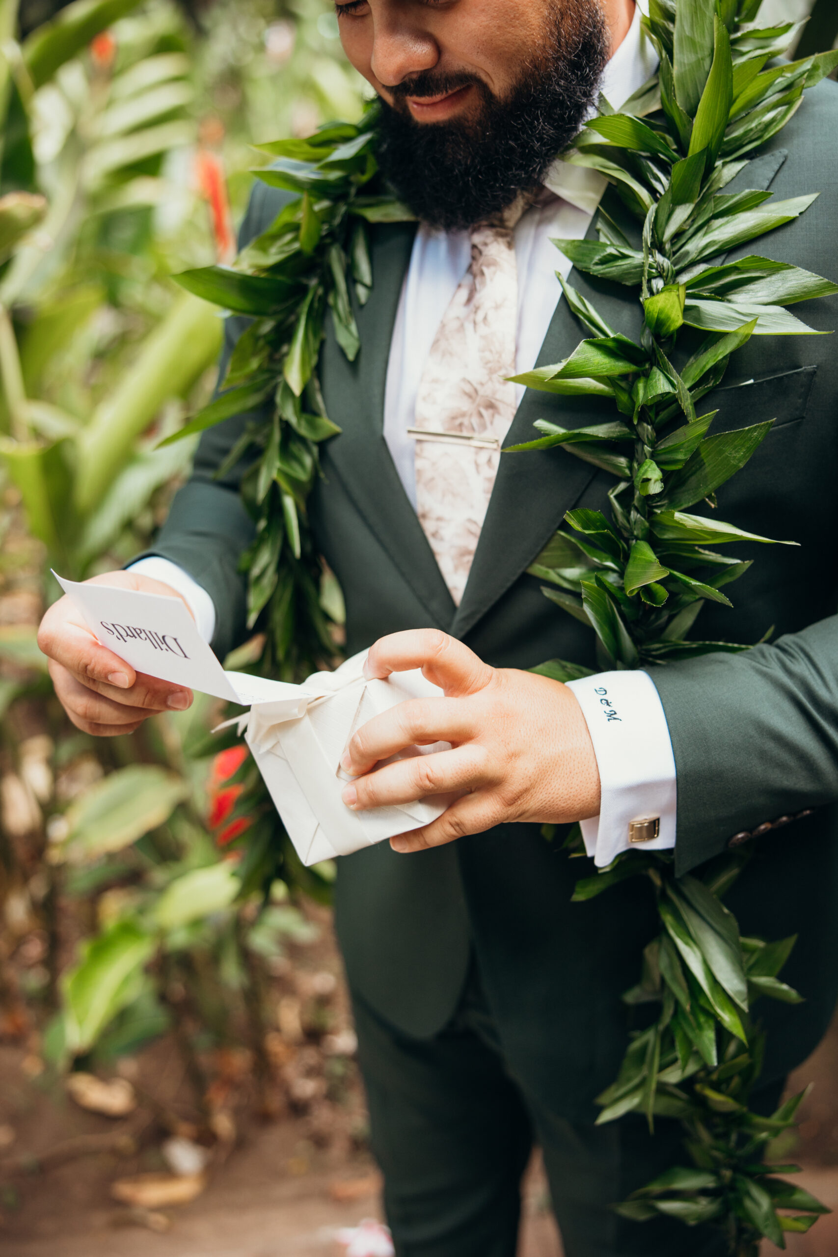 Groom opening a heartfelt wedding gift in a tropical garden, wearing a green suit and maile lei with “Forever” embroidered on his cuff.