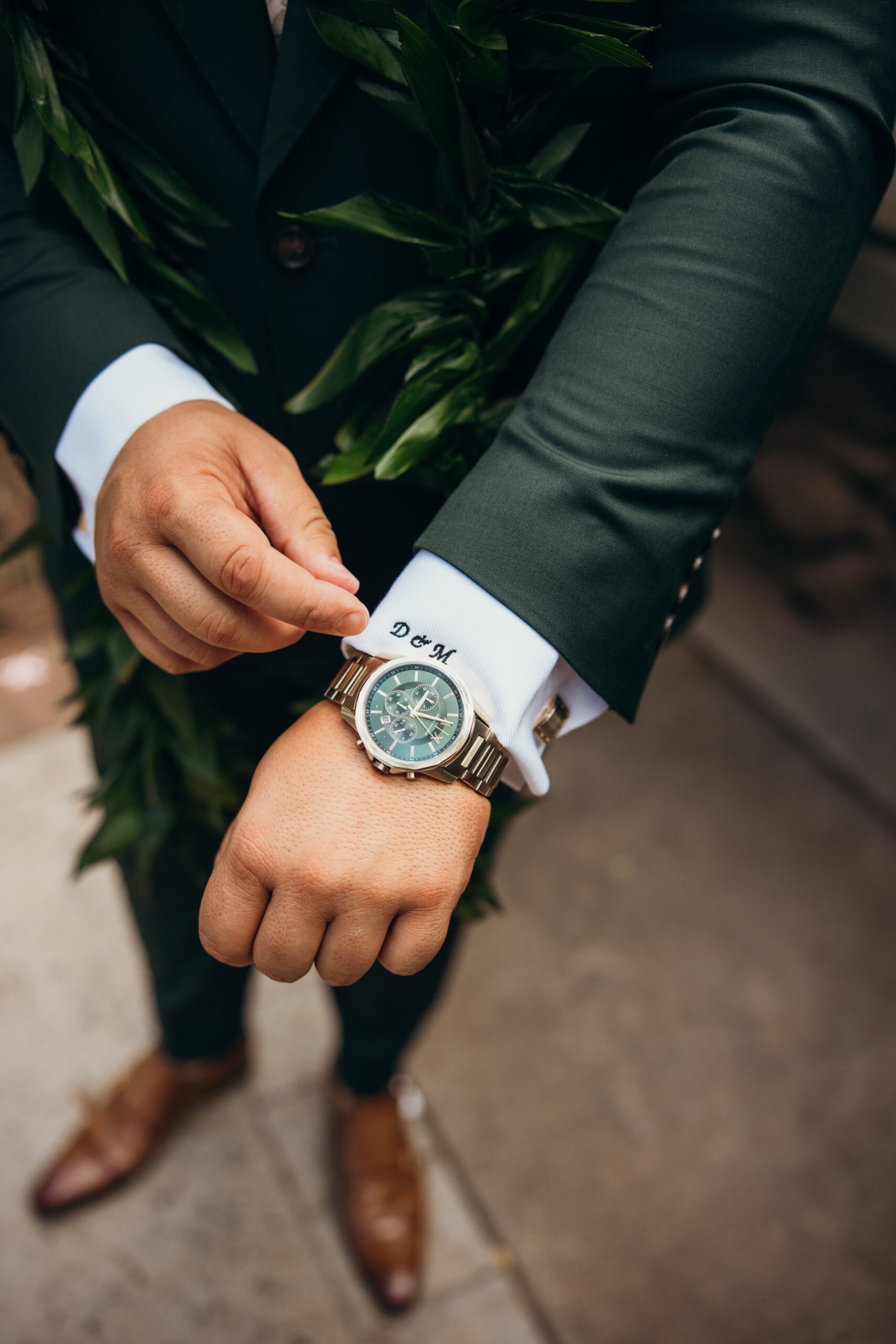 Close-up of the groom adjusting his cuff with embroidered initials and wearing a luxury watch, dressed in a deep green suit with a maile lei.