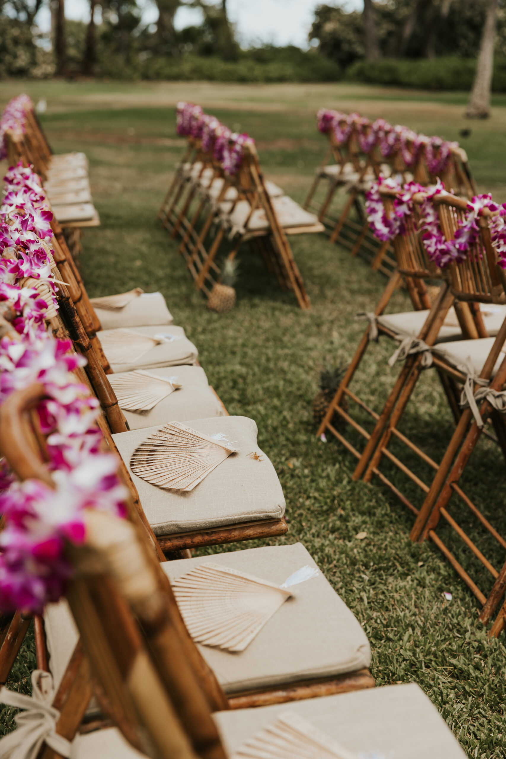 Bamboo ceremony chairs adorned with purple orchid leis and woven fans, ready for guests at a scenic Maui wedding venue.