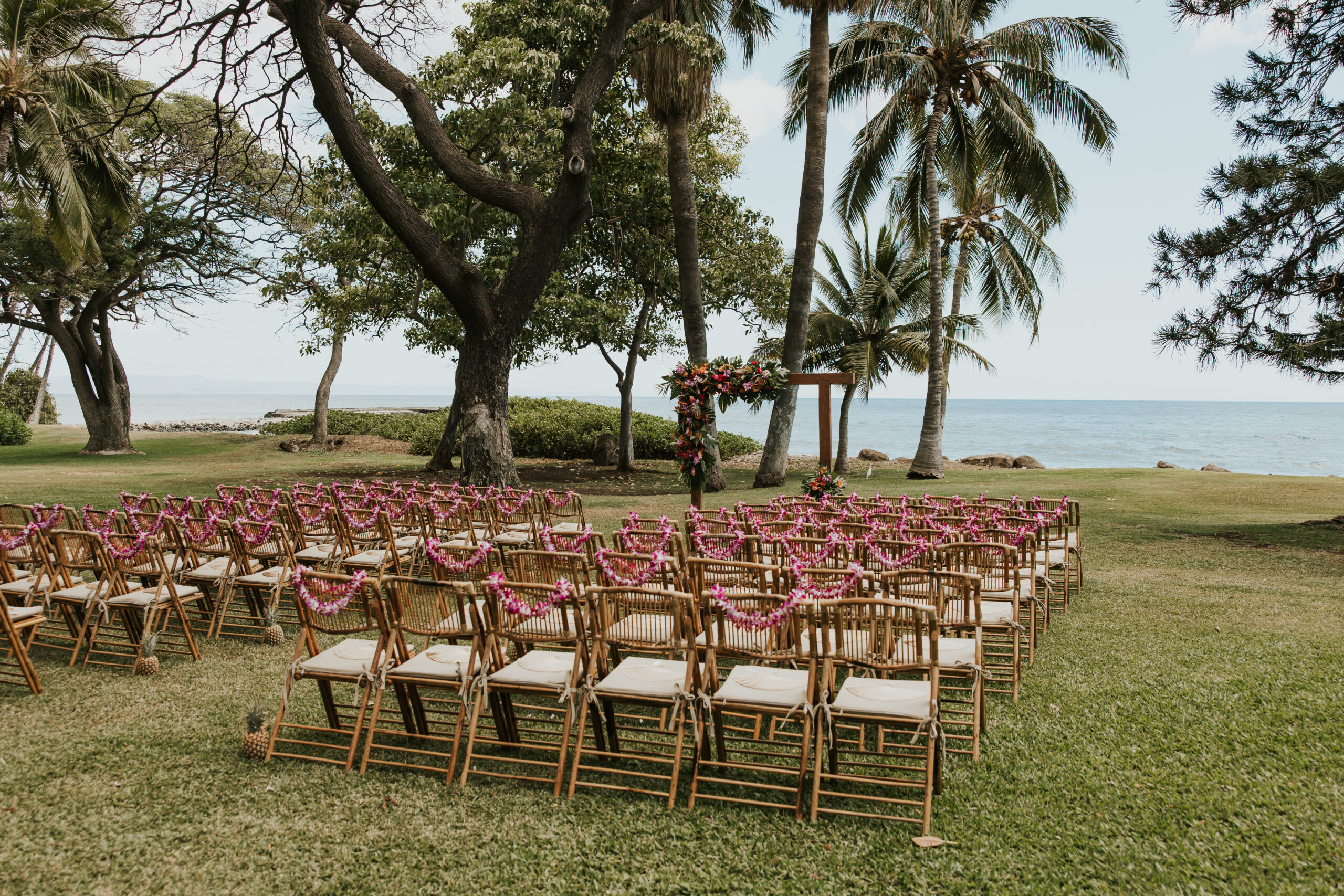 Oceanfront ceremony space with rows of bamboo chairs draped in orchid leis, set beneath towering palm trees at a Maui wedding venue.