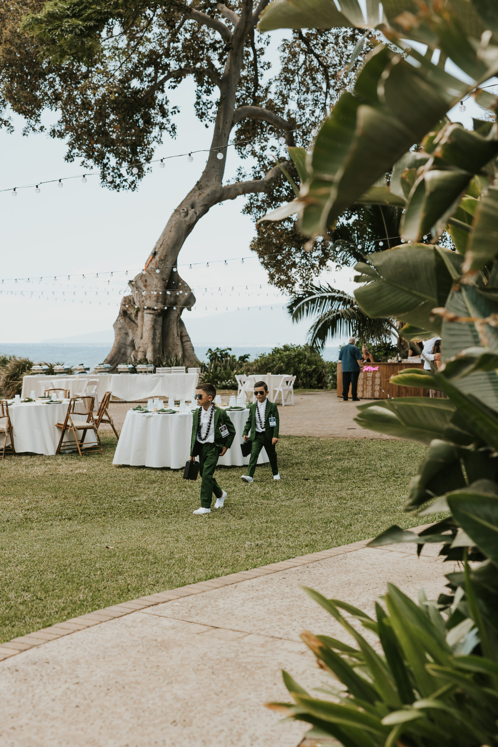 Two young ring bearers in green suits walking across a lawn with ocean views, helping set the scene at a relaxed Maui wedding venue.