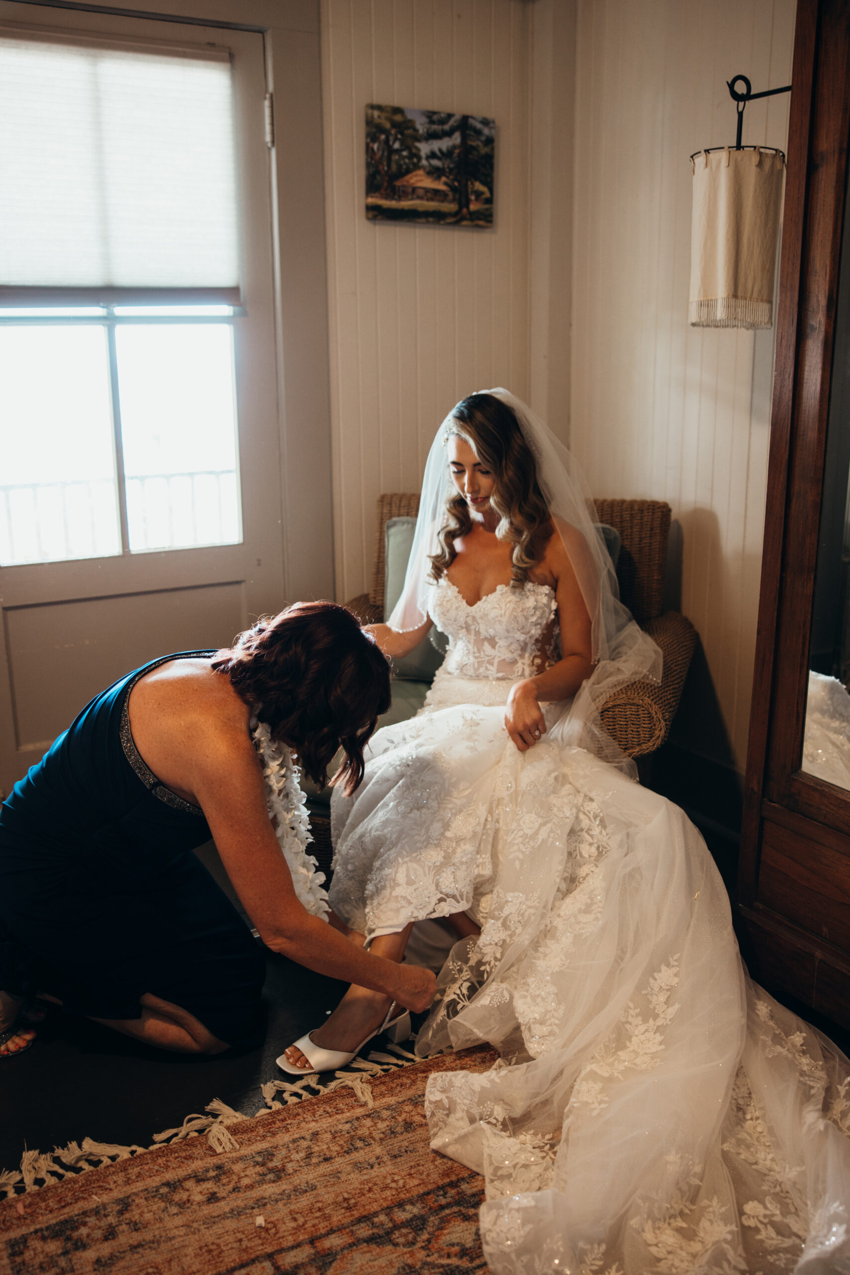 Mother of the bride kneels to fasten her daughter’s shoe as the bride sits in a wicker chair, glowing in her wedding gown.