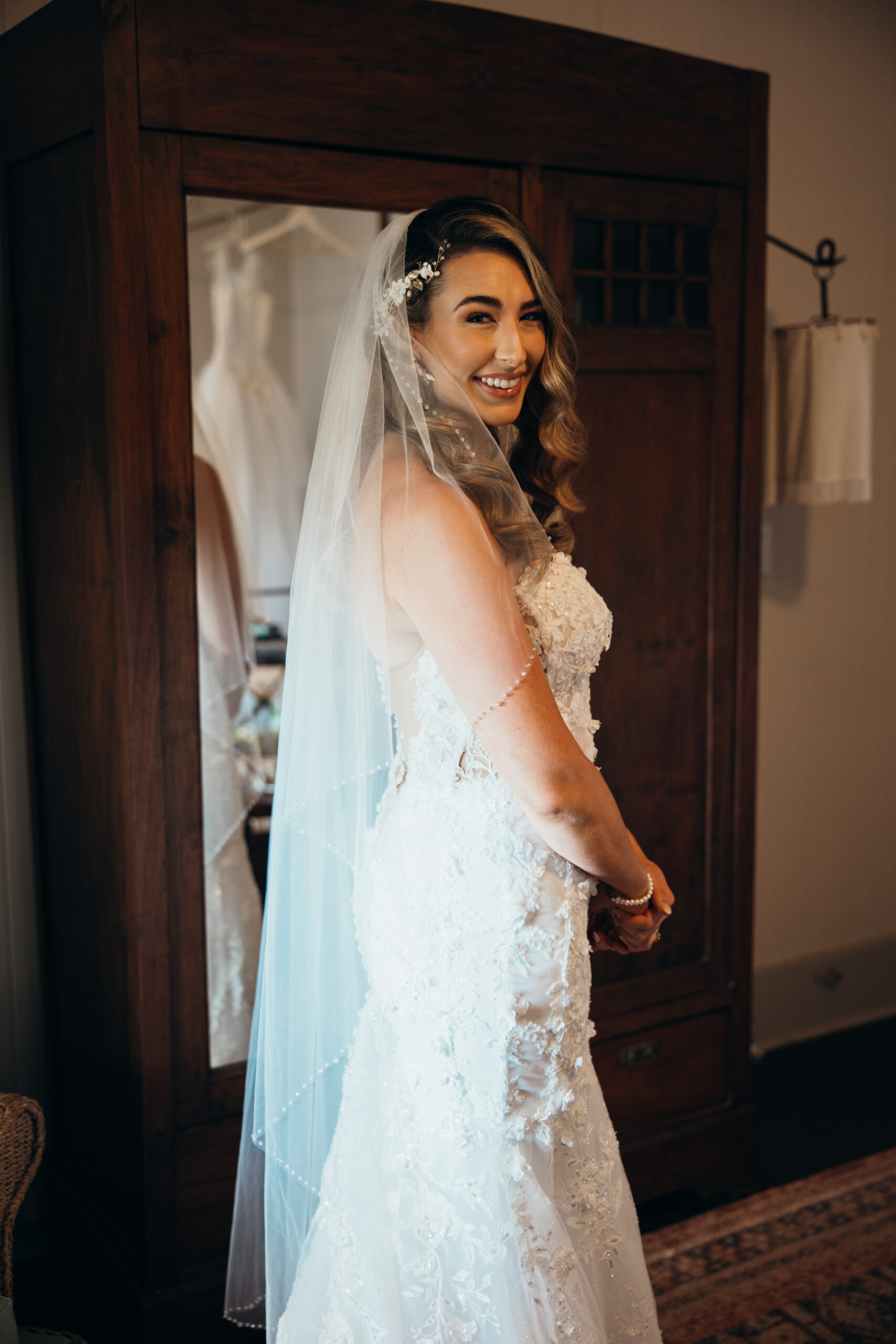 Radiant bride smiling in her lace wedding dress and veil while getting ready inside a cozy room filled with vintage touches.