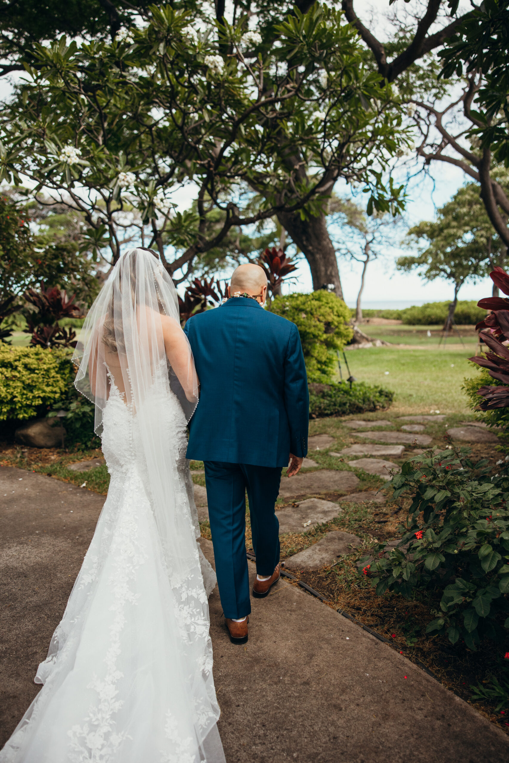Bride walks arm-in-arm with her father along a garden path, heading toward the ceremony space for her Maui wedding.