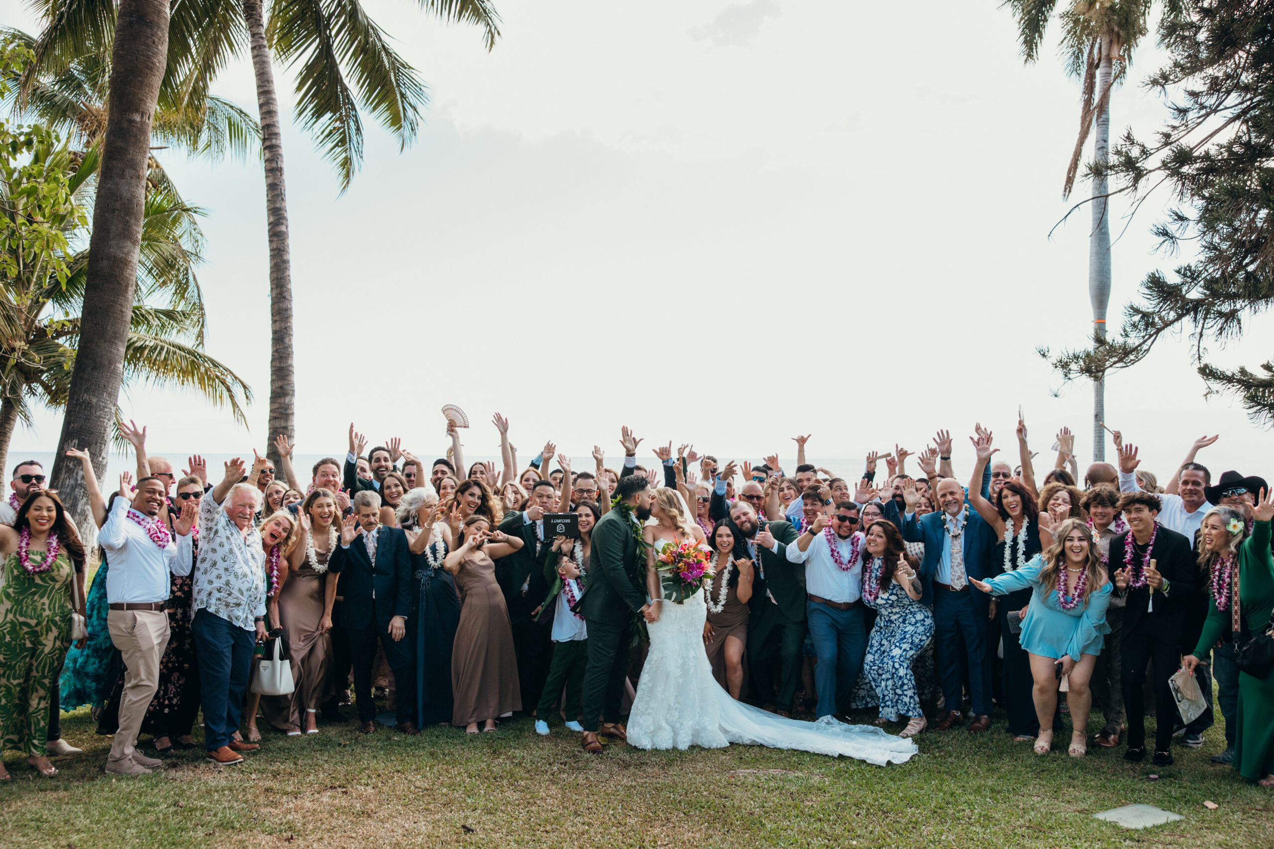 Bride and groom share a kiss while surrounded by their entire wedding party and guests cheering beneath the palms at a Maui wedding venue.