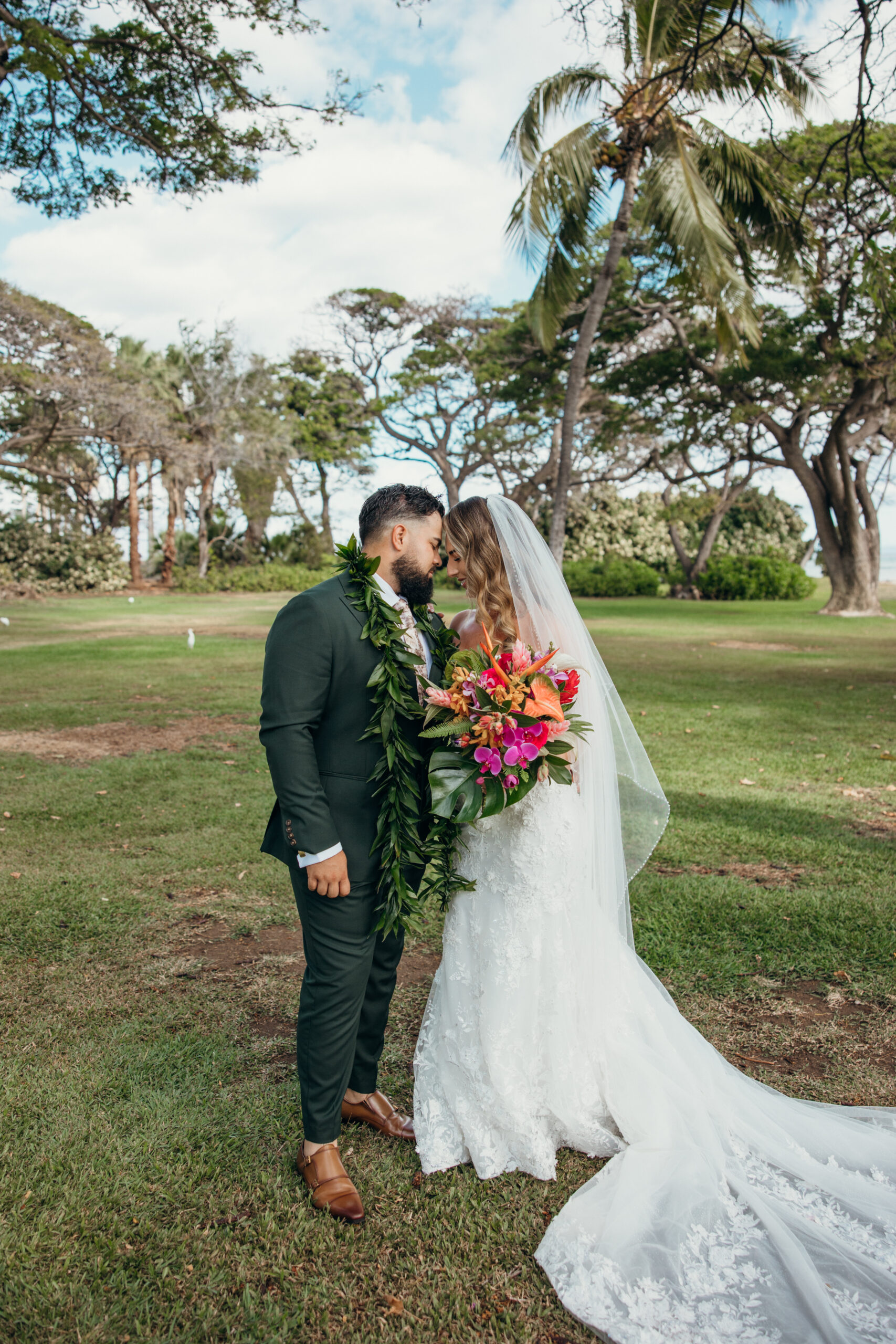 Bride and groom sharing a sweet forehead kiss under swaying palms, with her tropical bouquet adding a burst of color.