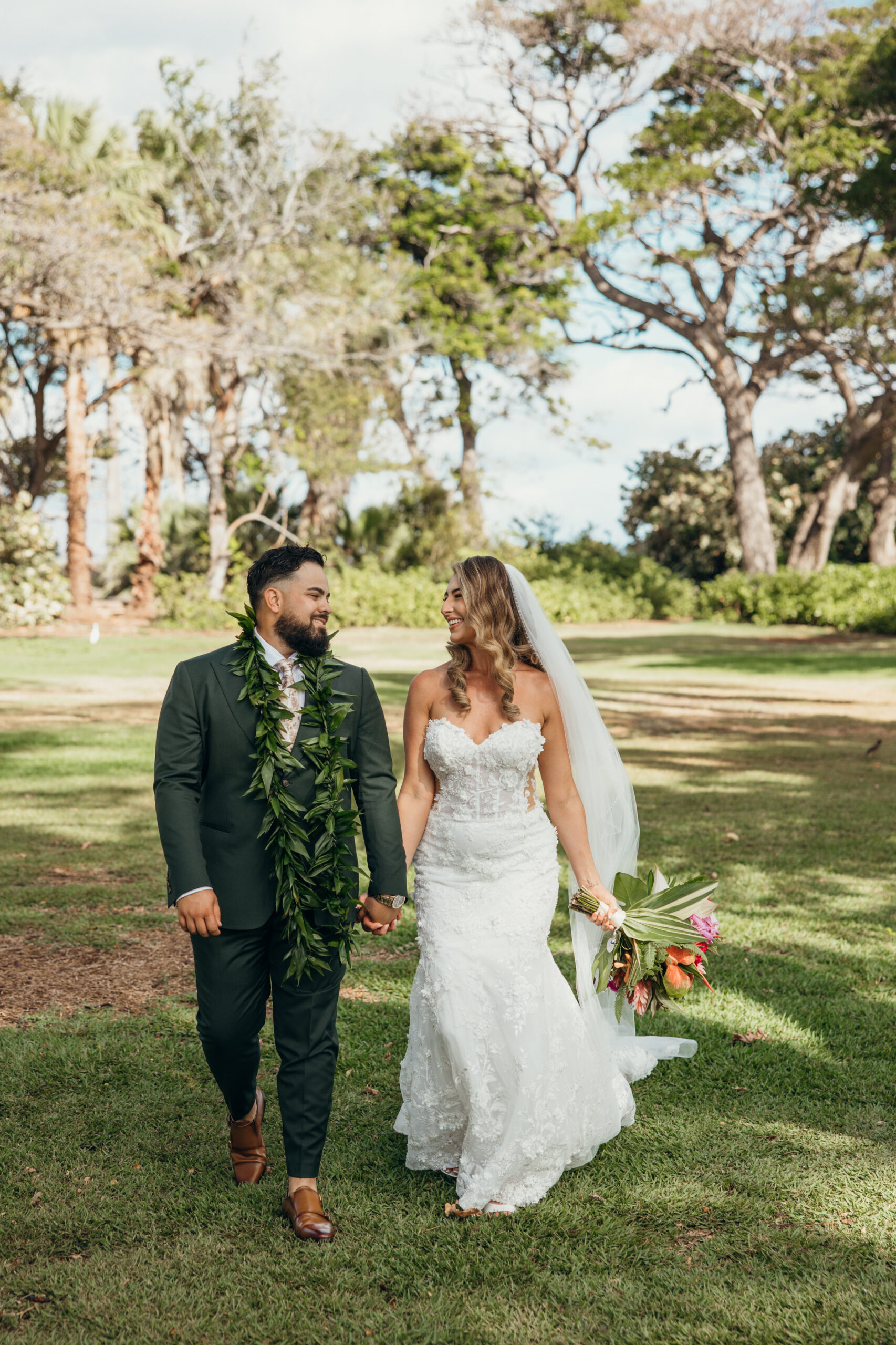 Bride and groom walk hand-in-hand across the lawn, smiling at each other after saying “I do” at a peaceful Maui wedding venue.