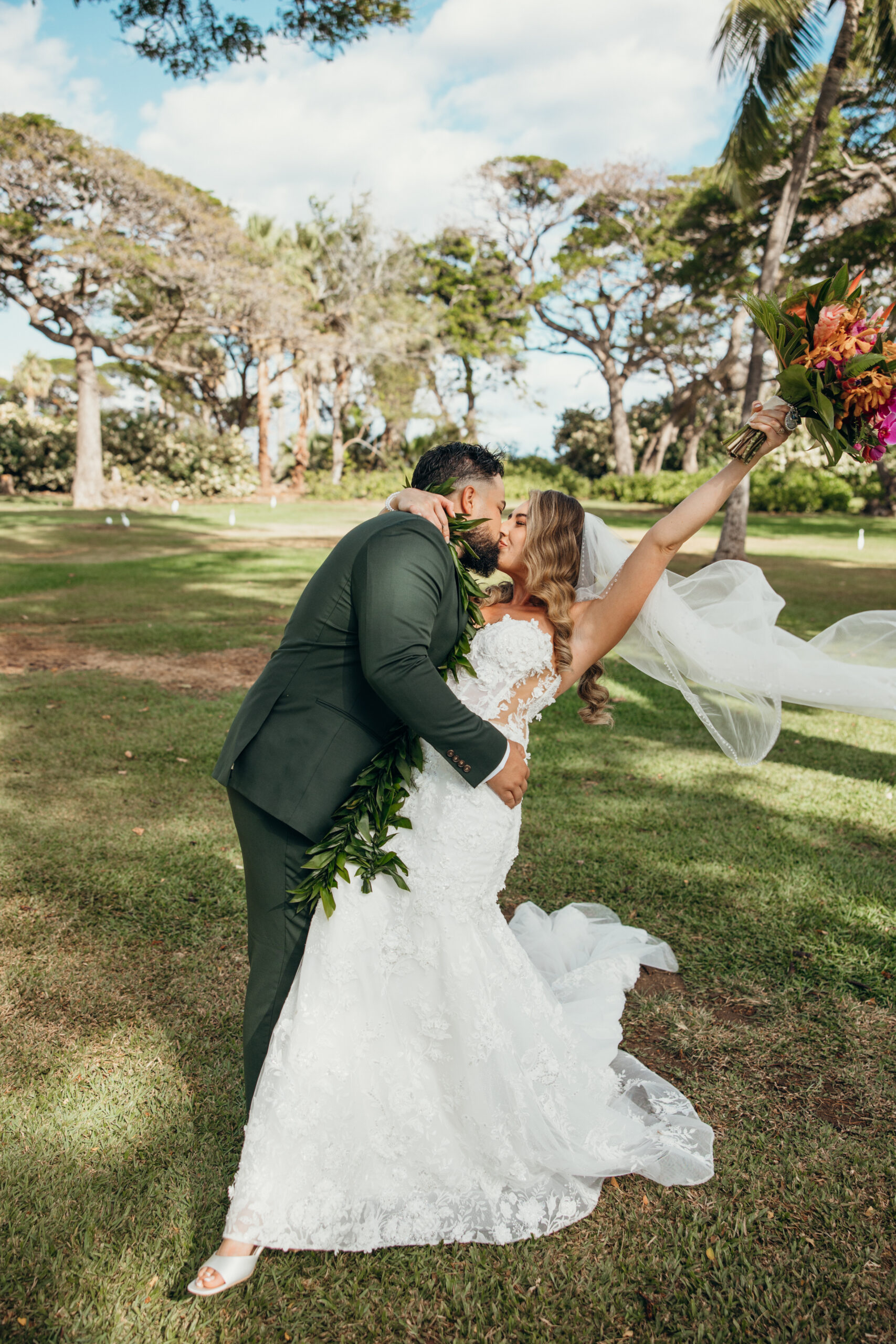 Groom dips his bride dramatically for a kiss, her veil flowing and bouquet lifted high during golden hour at a Maui wedding venue.