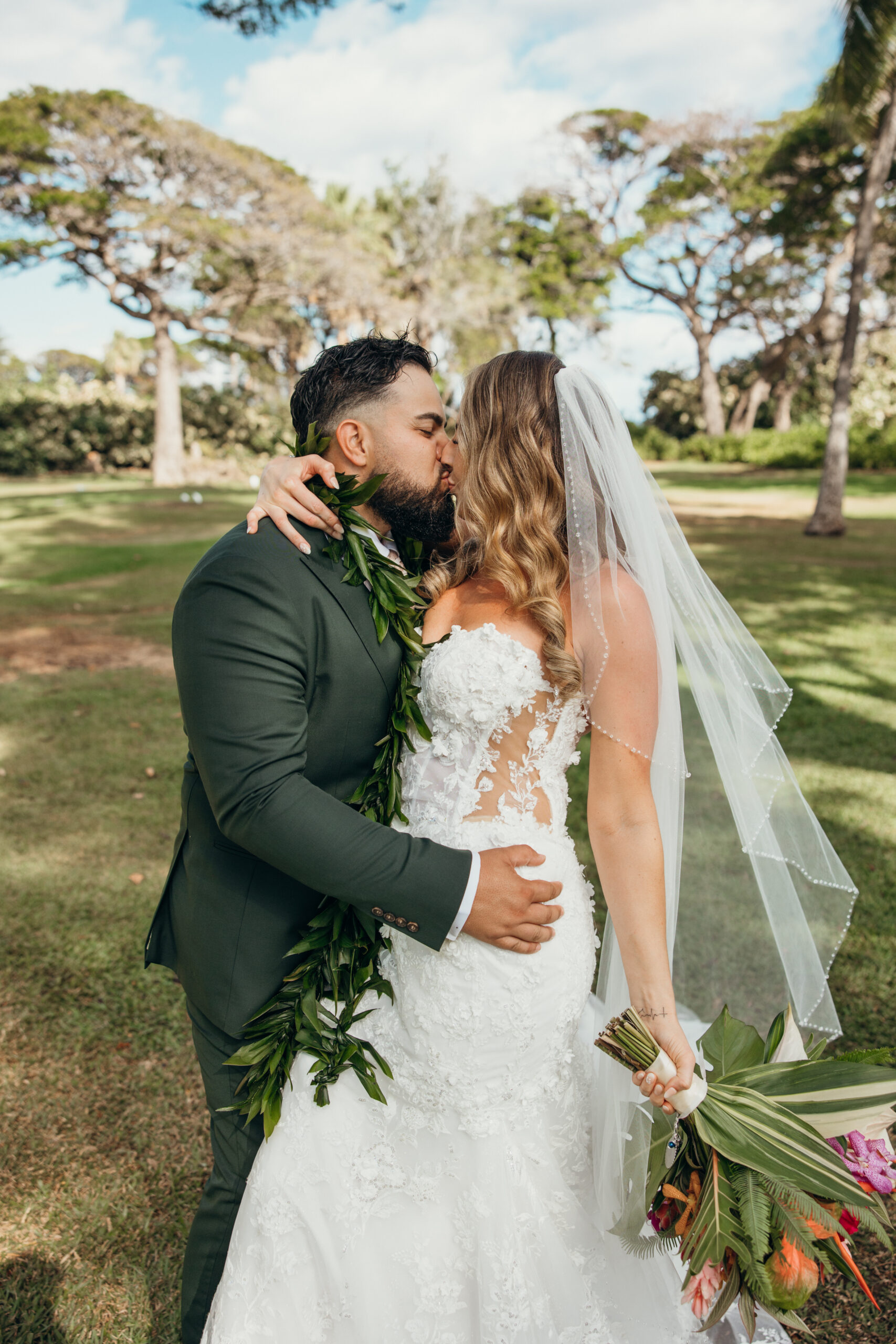 Just married kiss between the newlyweds, with her tropical bouquet in hand and his maile lei flowing in the breeze.