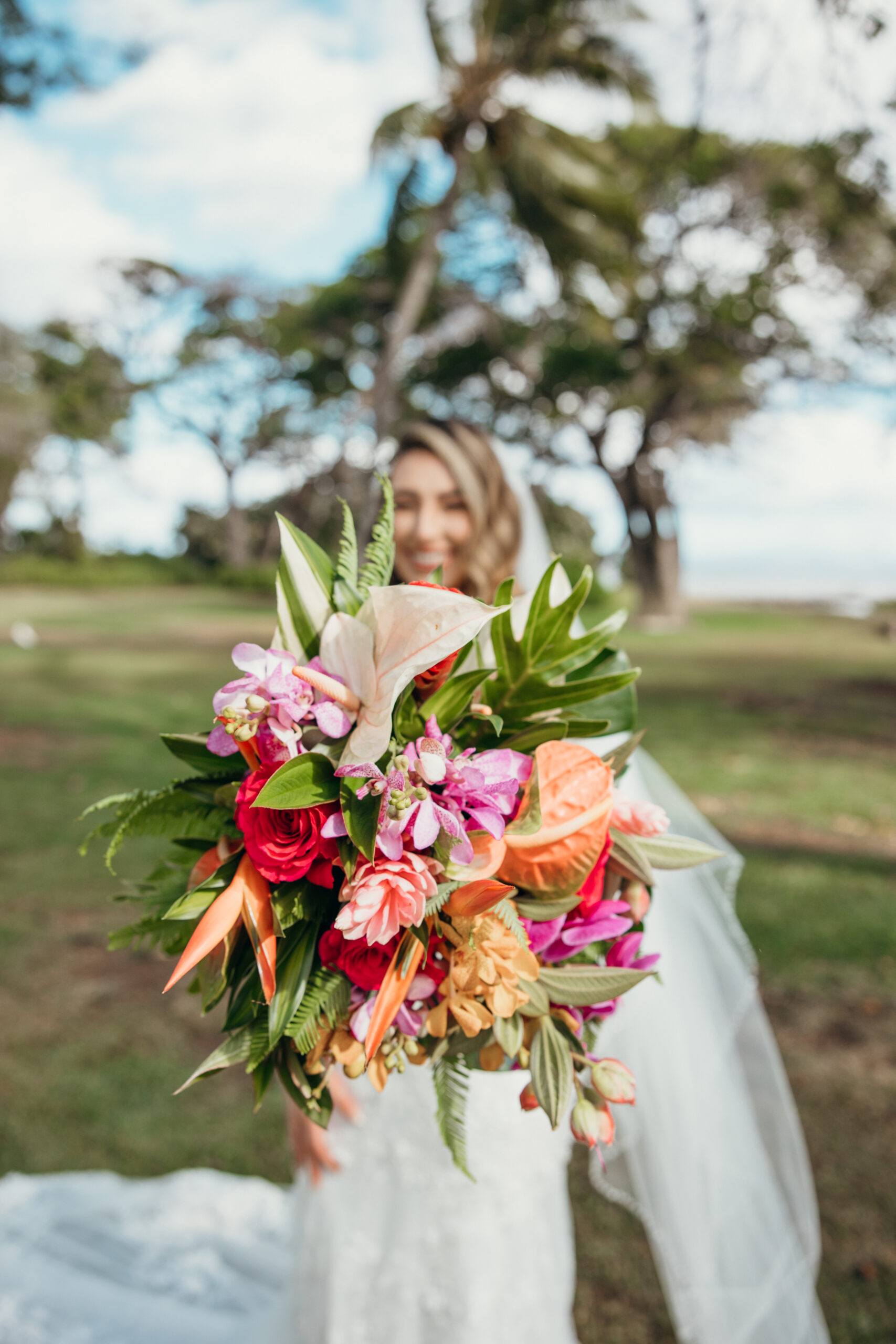 Colorful tropical bouquet held out toward the camera, with the smiling bride softly blurred in the background.