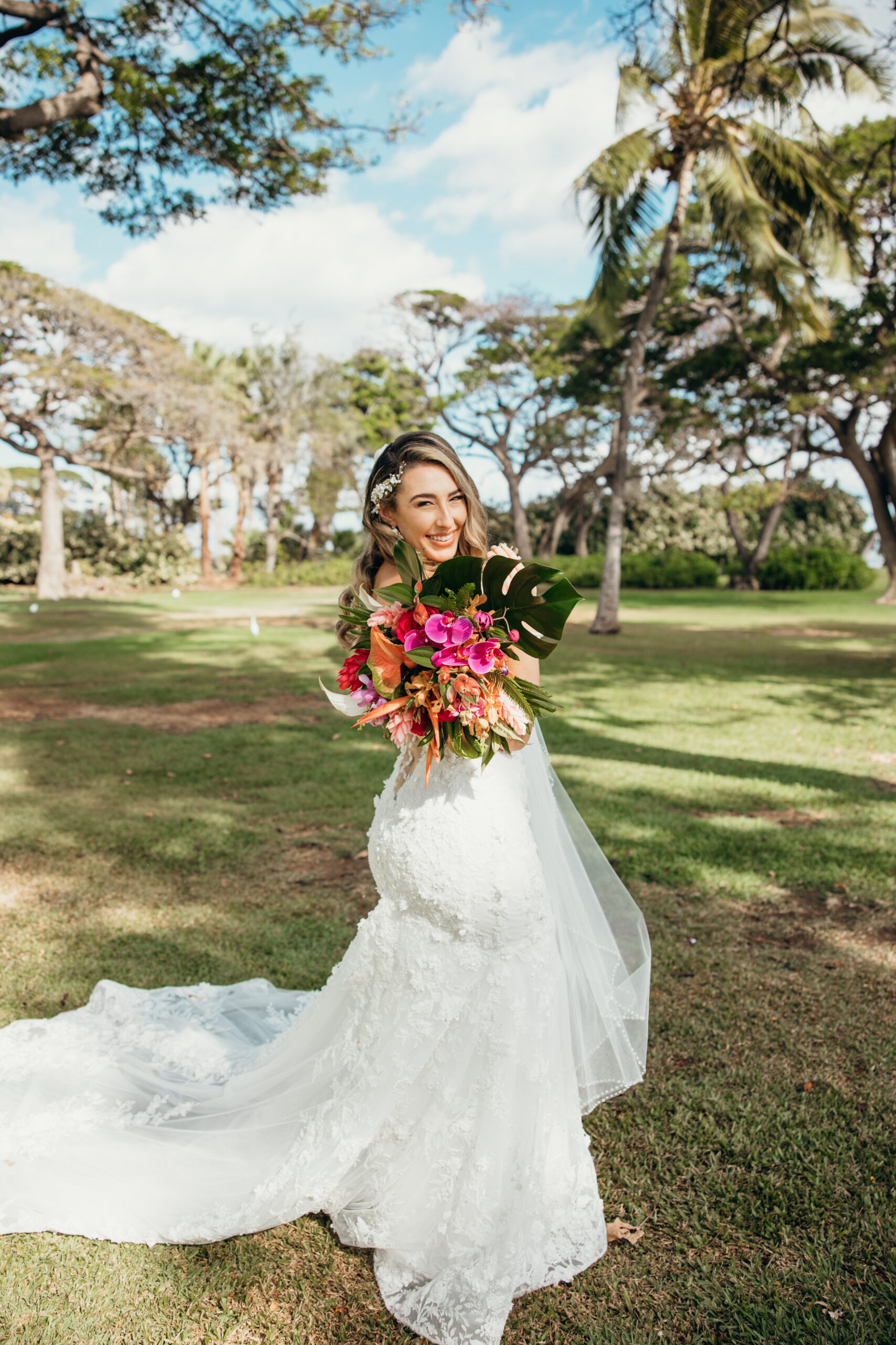 Bride smiling brightly, holding her vibrant tropical bouquet and standing beneath the palms at a peaceful Maui wedding venue.