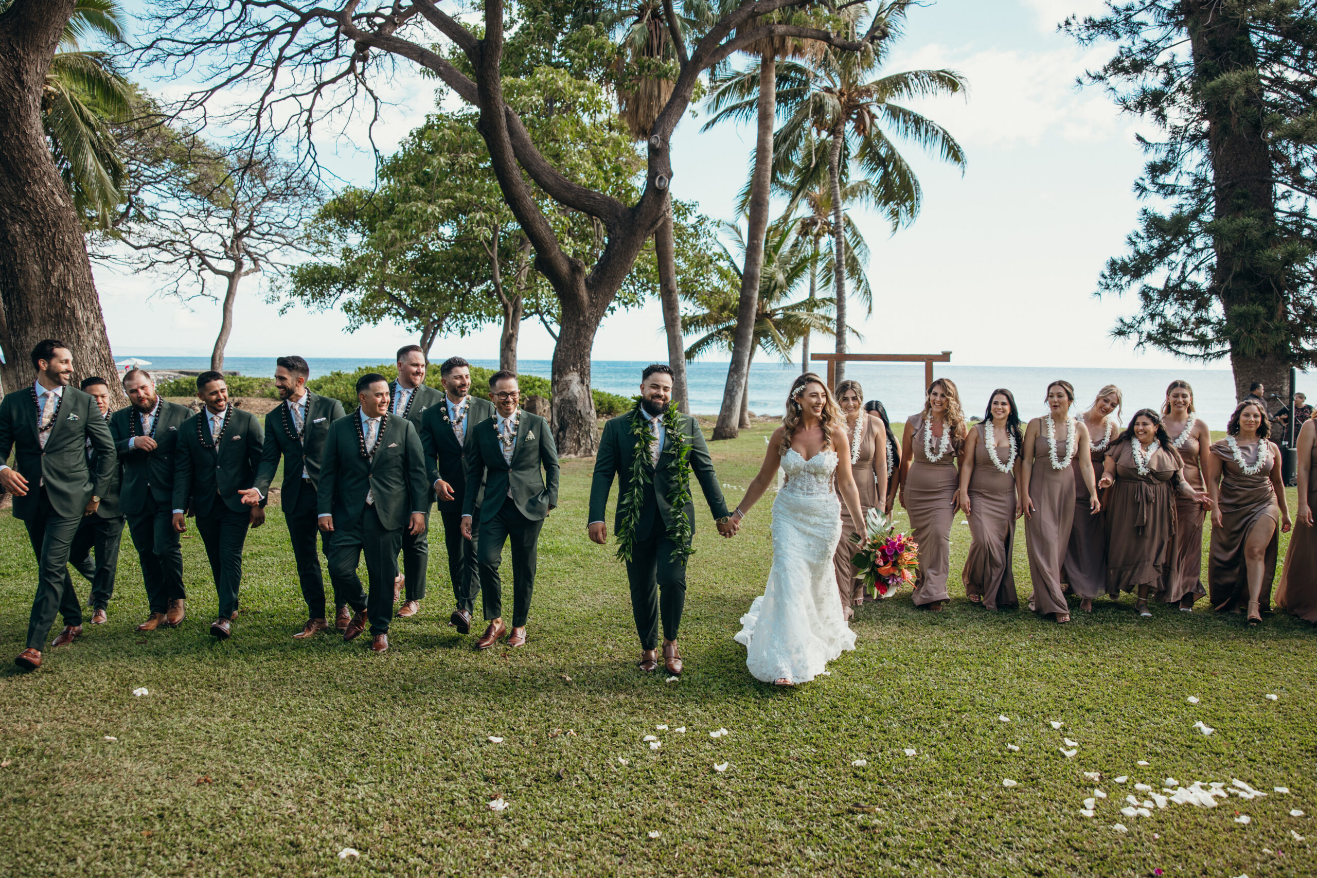 Bride and groom walking hand in hand with their full wedding party at a tropical Maui wedding venue, surrounded by palm trees and ocean views.