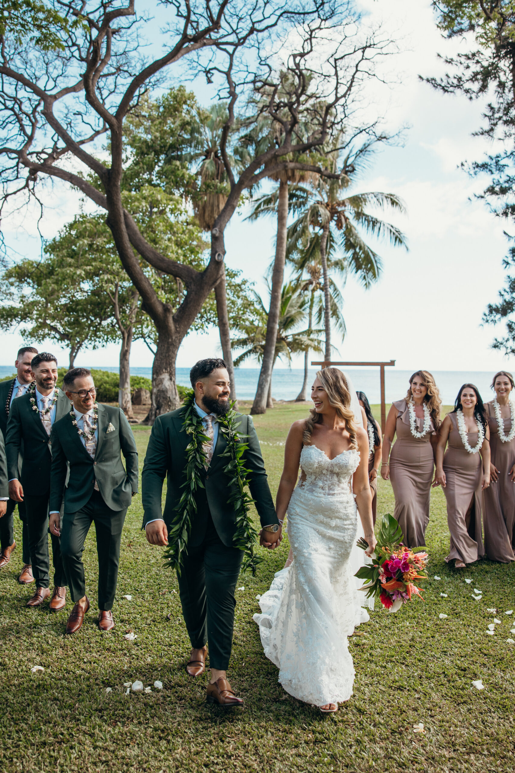 Bride and groom walk hand in hand, smiling at each other with their wedding party following behind at a lush Maui wedding venue.