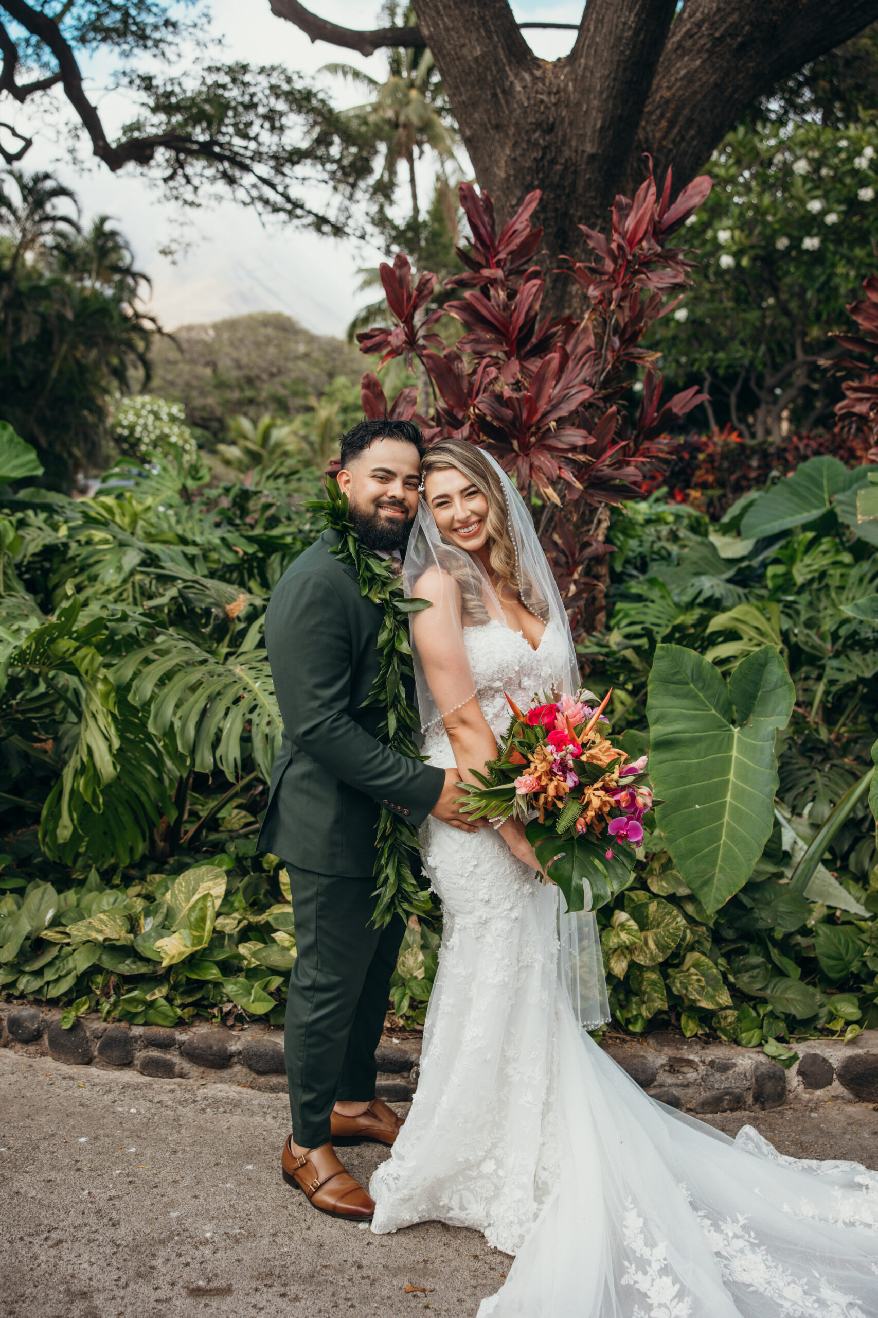 Bride and groom pose joyfully in front of lush tropical foliage, holding a bold tropical bouquet at their Maui wedding venue.