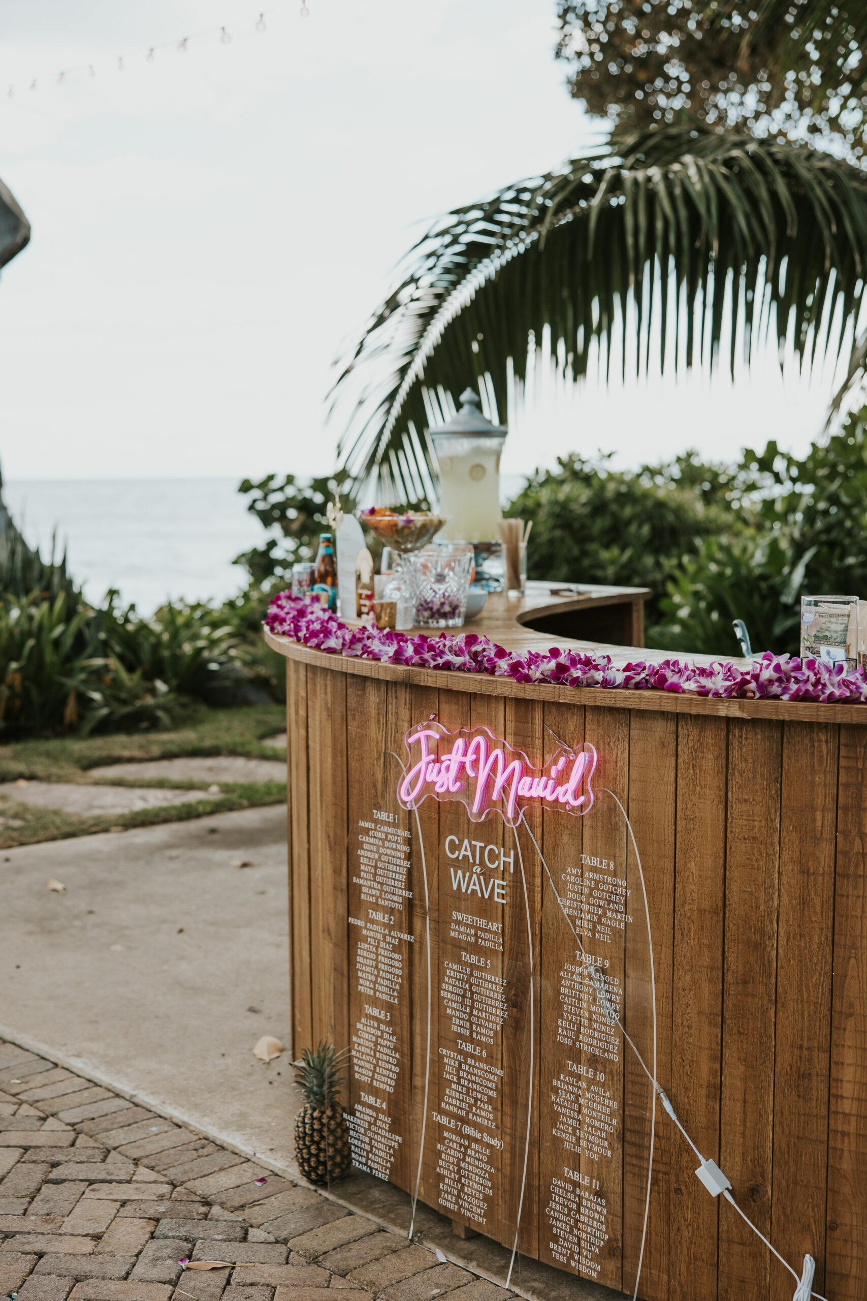 Tropical drink and escort card station at a Maui wedding venue, featuring a “Just Mauied” neon sign and flower leis with an ocean view.
