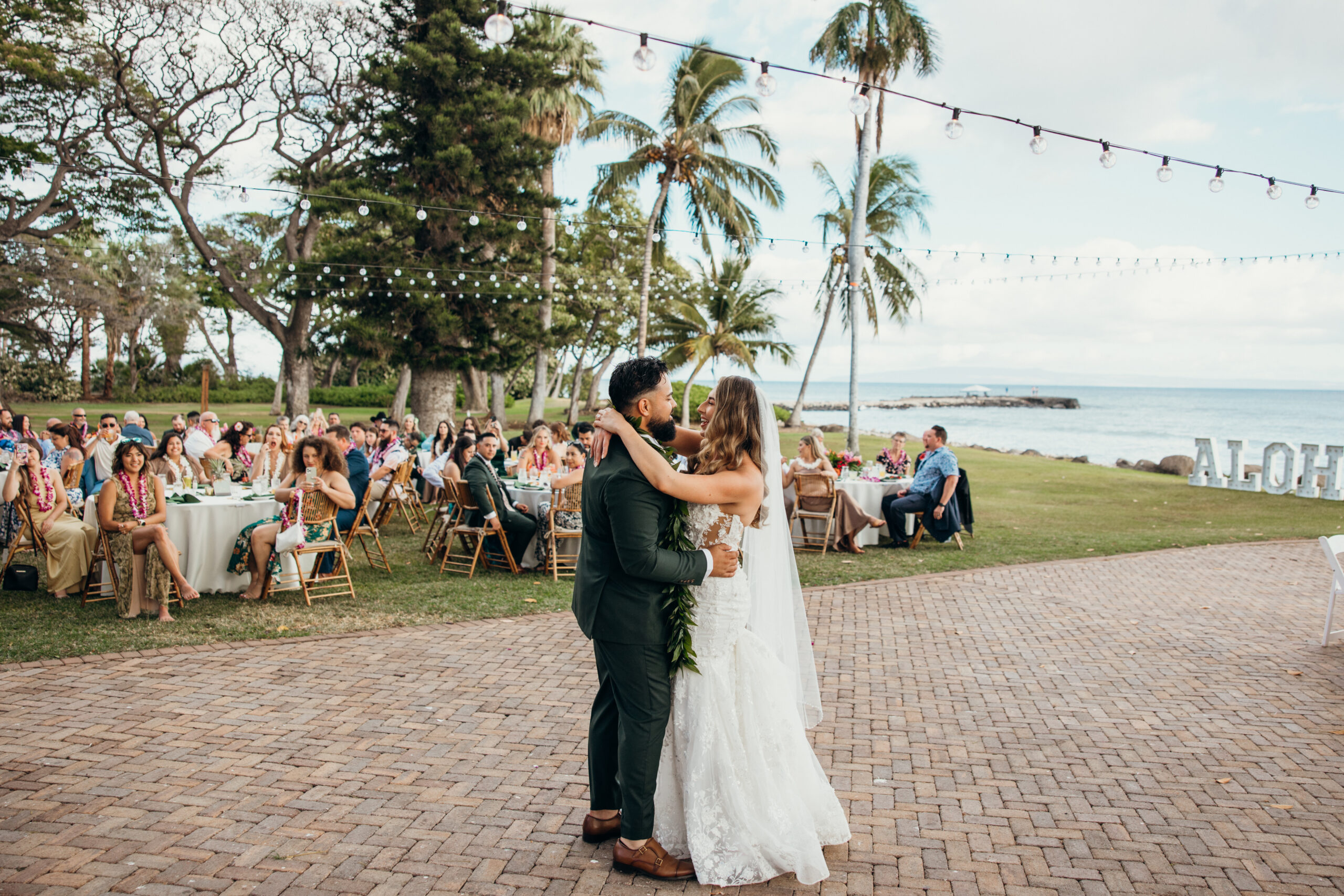 Newlyweds share their first dance under string lights with the ocean and palm trees in the background at a lively Maui wedding venue.