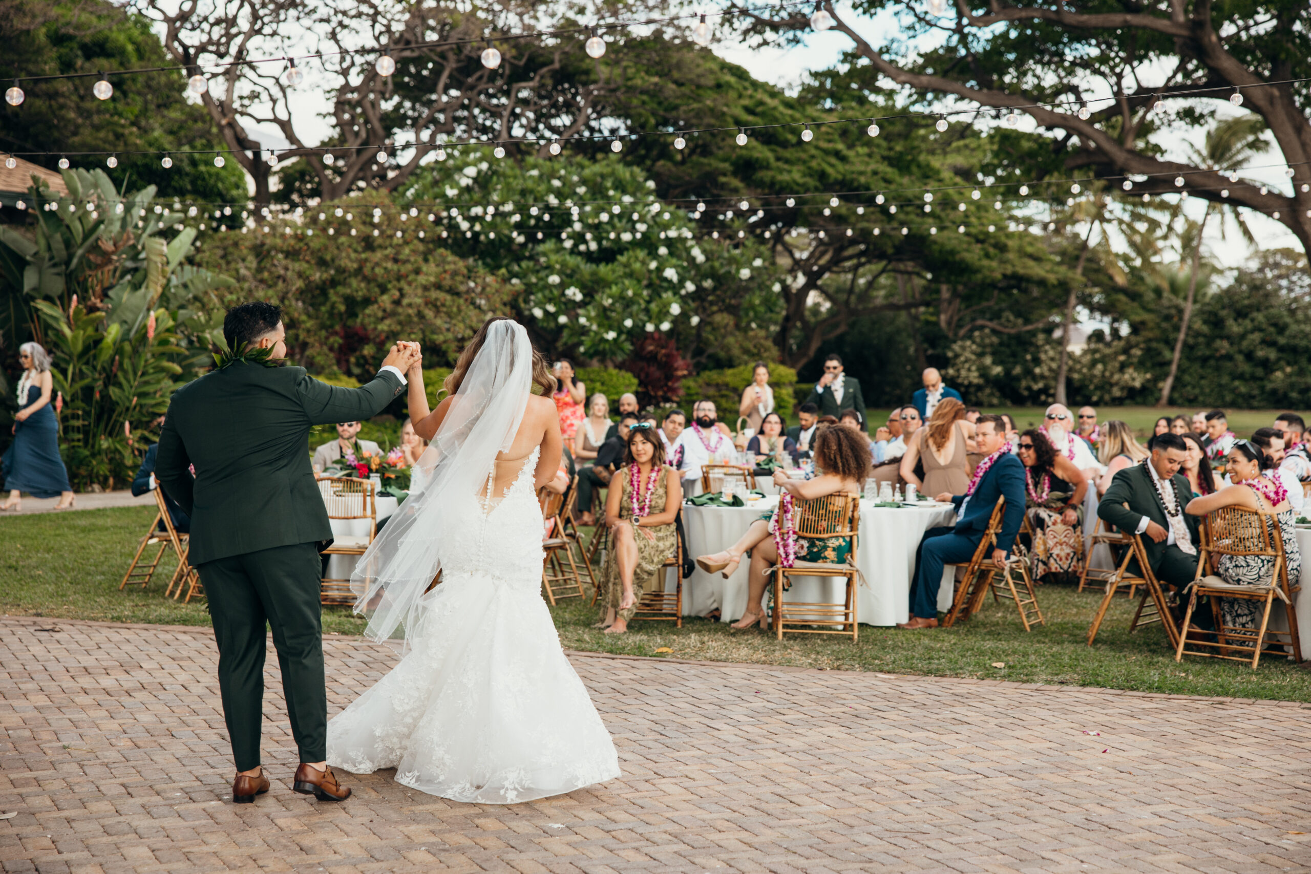 Bride and groom twirl into their reception as guests cheer beneath string lights at a tropical Maui wedding venue.