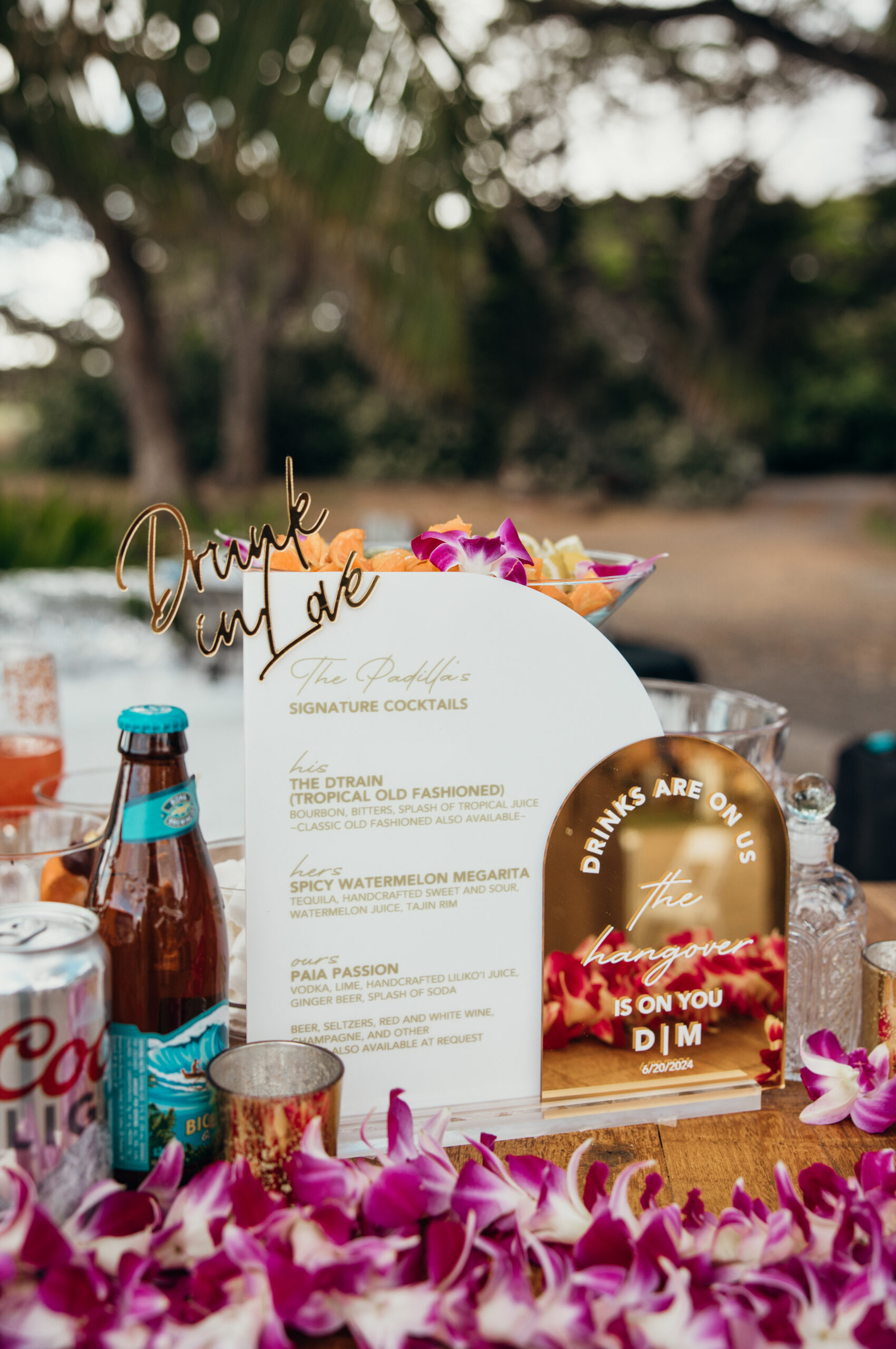 Signature cocktail menu and bar setup decorated with orchids, tropical drinks, and playful signage at a fun outdoor Maui wedding.