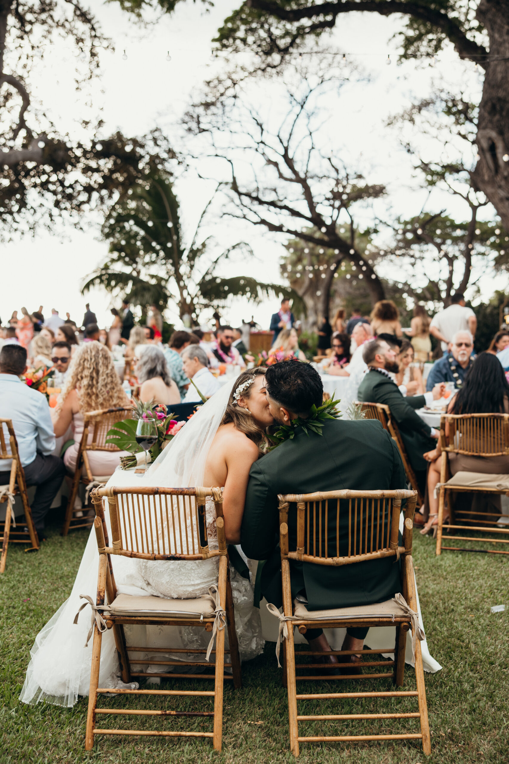 Bride and groom sneak a kiss while seated at their sweetheart table, surrounded by guests enjoying the celebration at a Maui wedding venue.