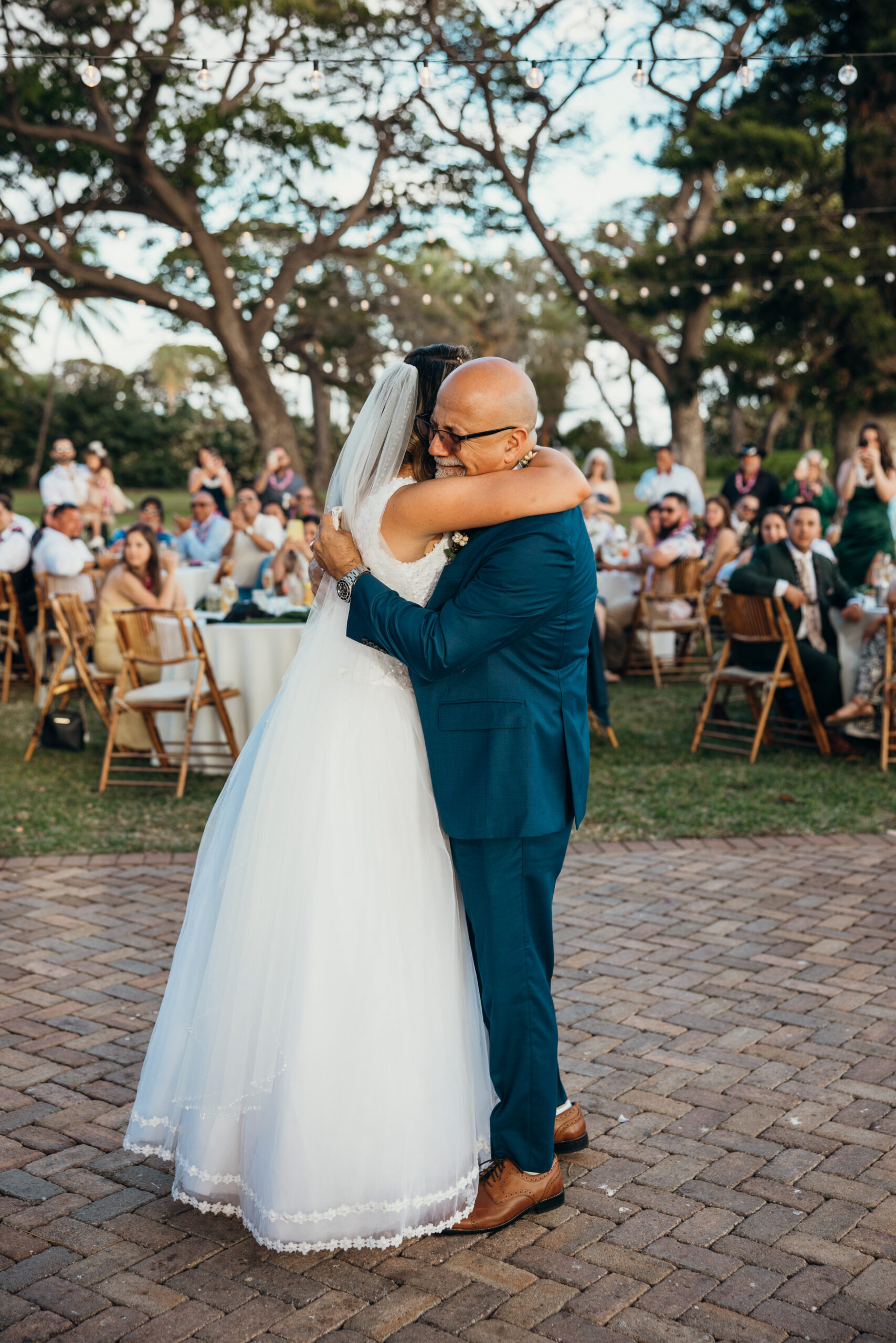 Father-daughter dance captured in a joyful embrace, with guests watching on under the trees at this heartfelt Maui reception.