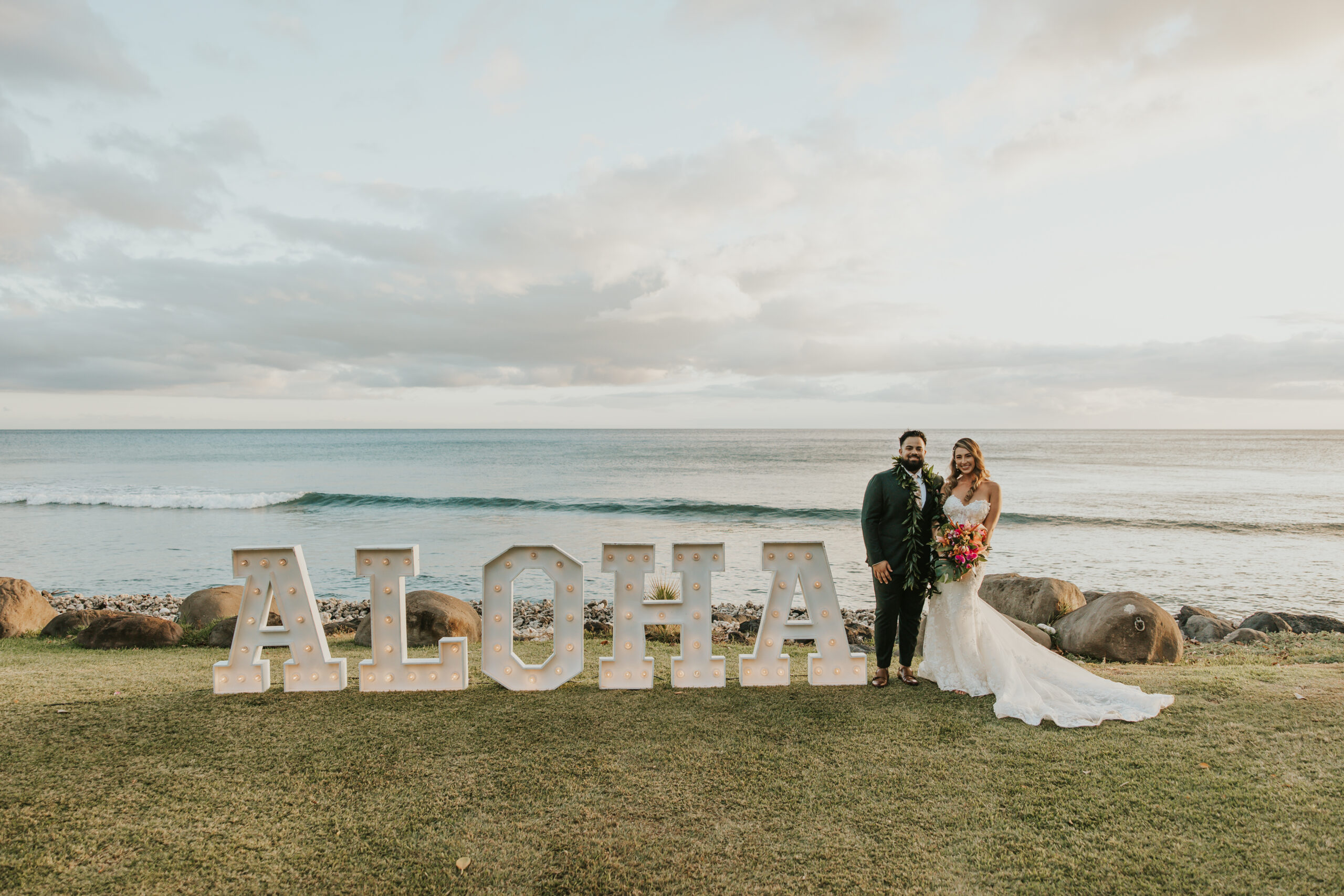 Couple poses in front of giant lit “ALOHA” letters by the ocean, a perfect golden hour moment at their Maui wedding venue.