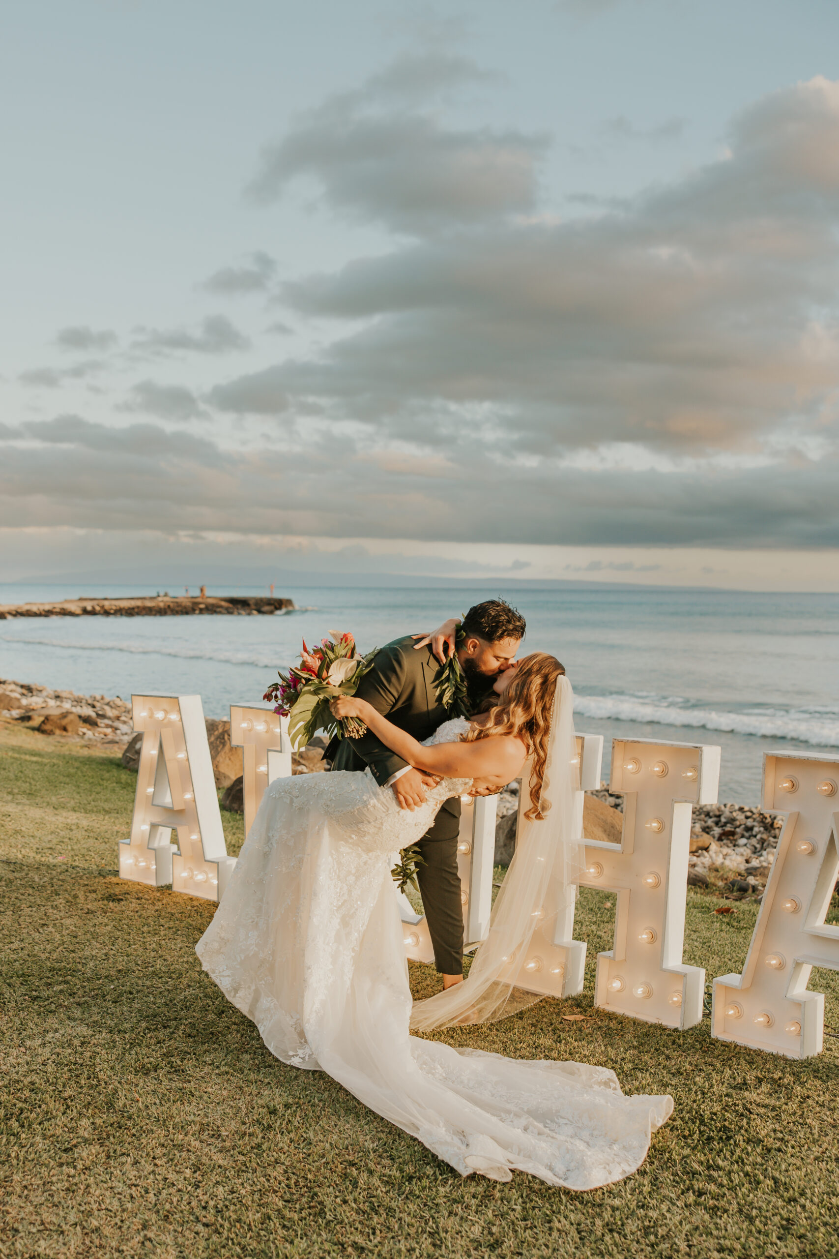 Groom dips bride for a romantic sunset kiss near the ocean, with giant “ALOHA” marquee letters glowing behind them at a Maui wedding venue.