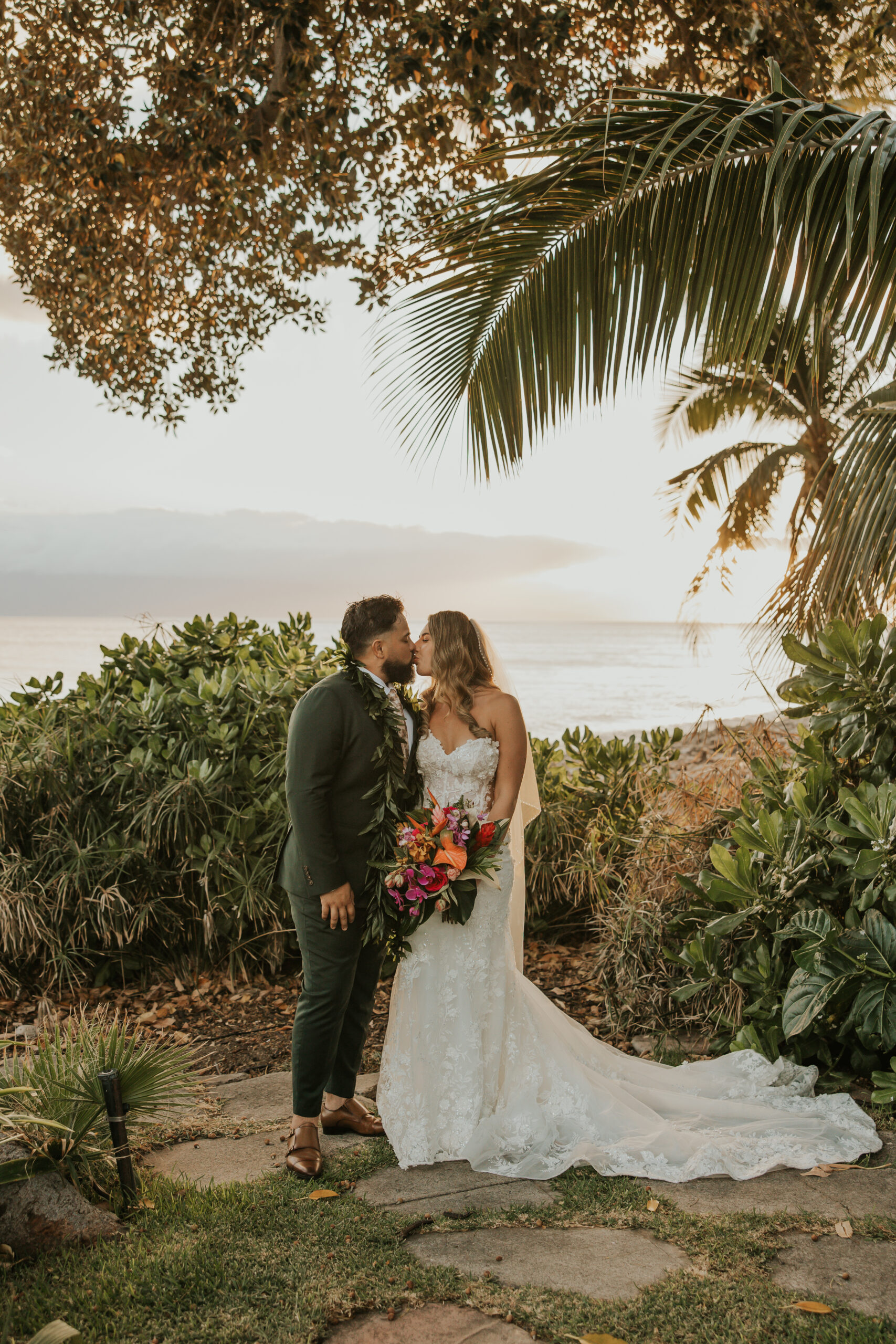 Bride and groom kiss beneath lush greenery and palm trees with the Maui coastline glowing in the sunset behind them.